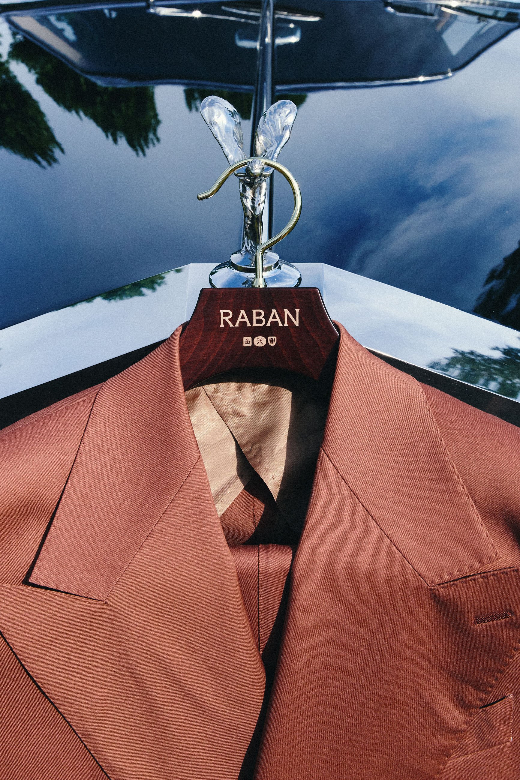Close-up of a clothing hanger with a pink blazer and a pink shirt hanging on it, with a hooded cape behind them, and a reflective surface showing trees in the background.