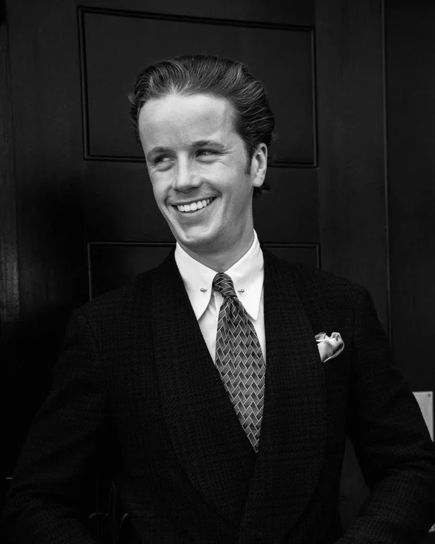 A young man with slicked-back hair, dressed in a dark suit, white shirt, patterned tie, and pocket square, smiling and looking to his right in front of a wooden door.