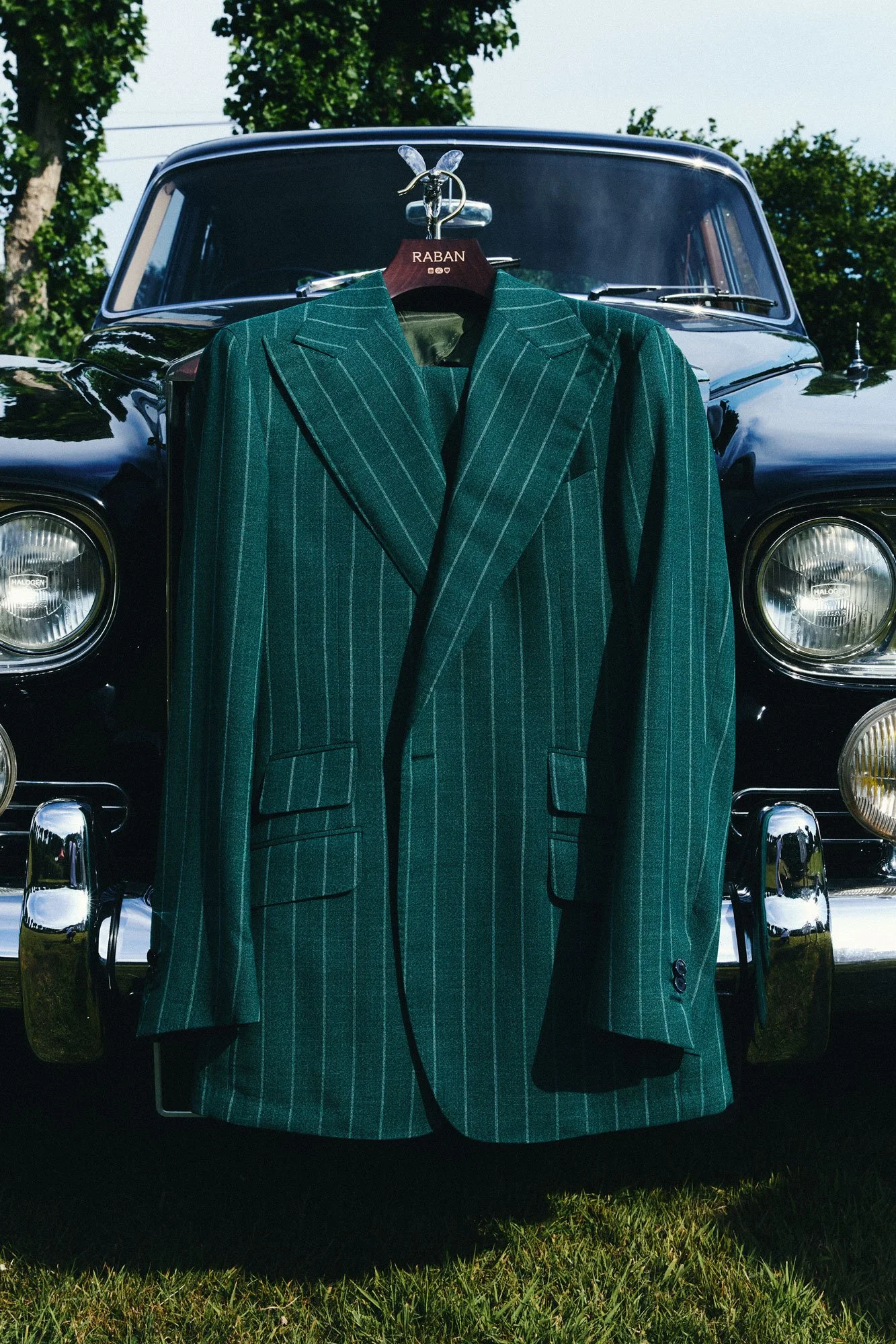 Green striped suit hanging on a hanger in front of a black car with trees in the background.