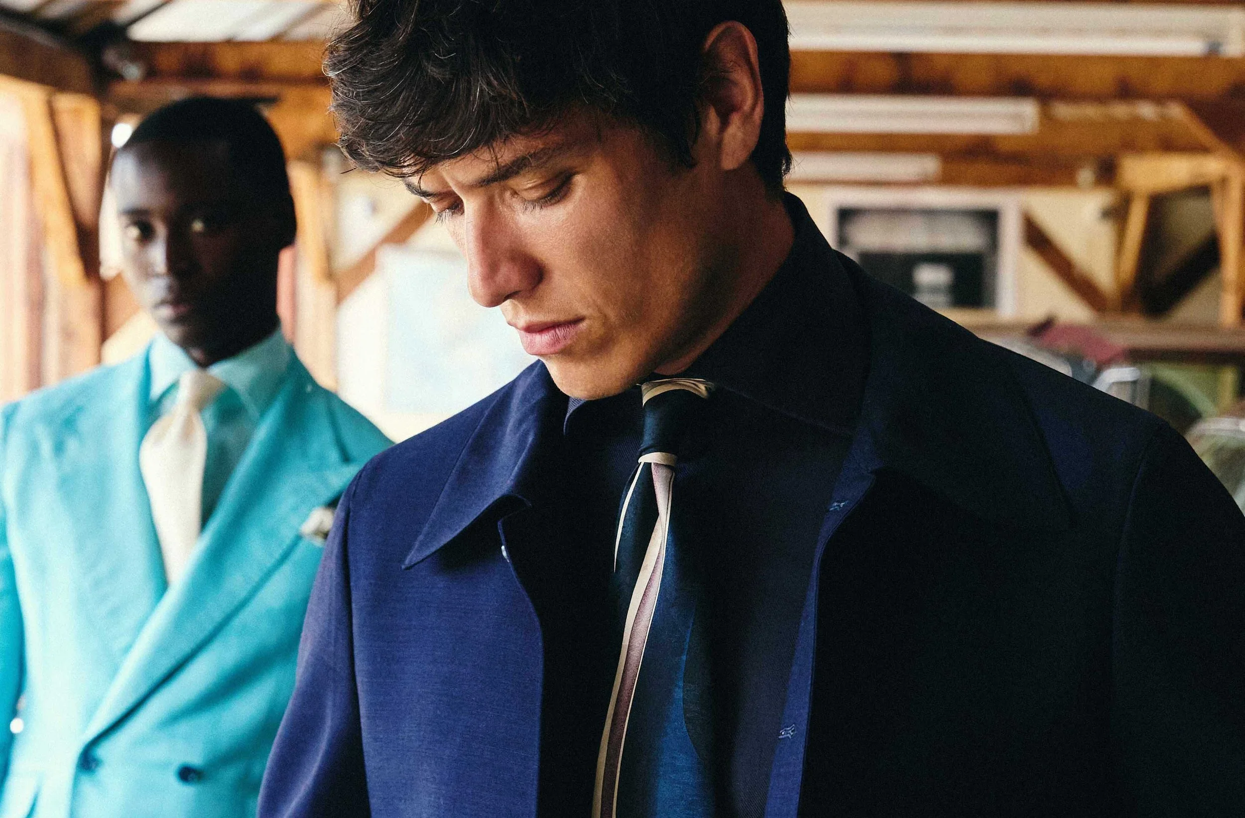 Young man in a dark blue suit with a striped tie looking down, standing indoors with a woman in turquoise clothing in the background.