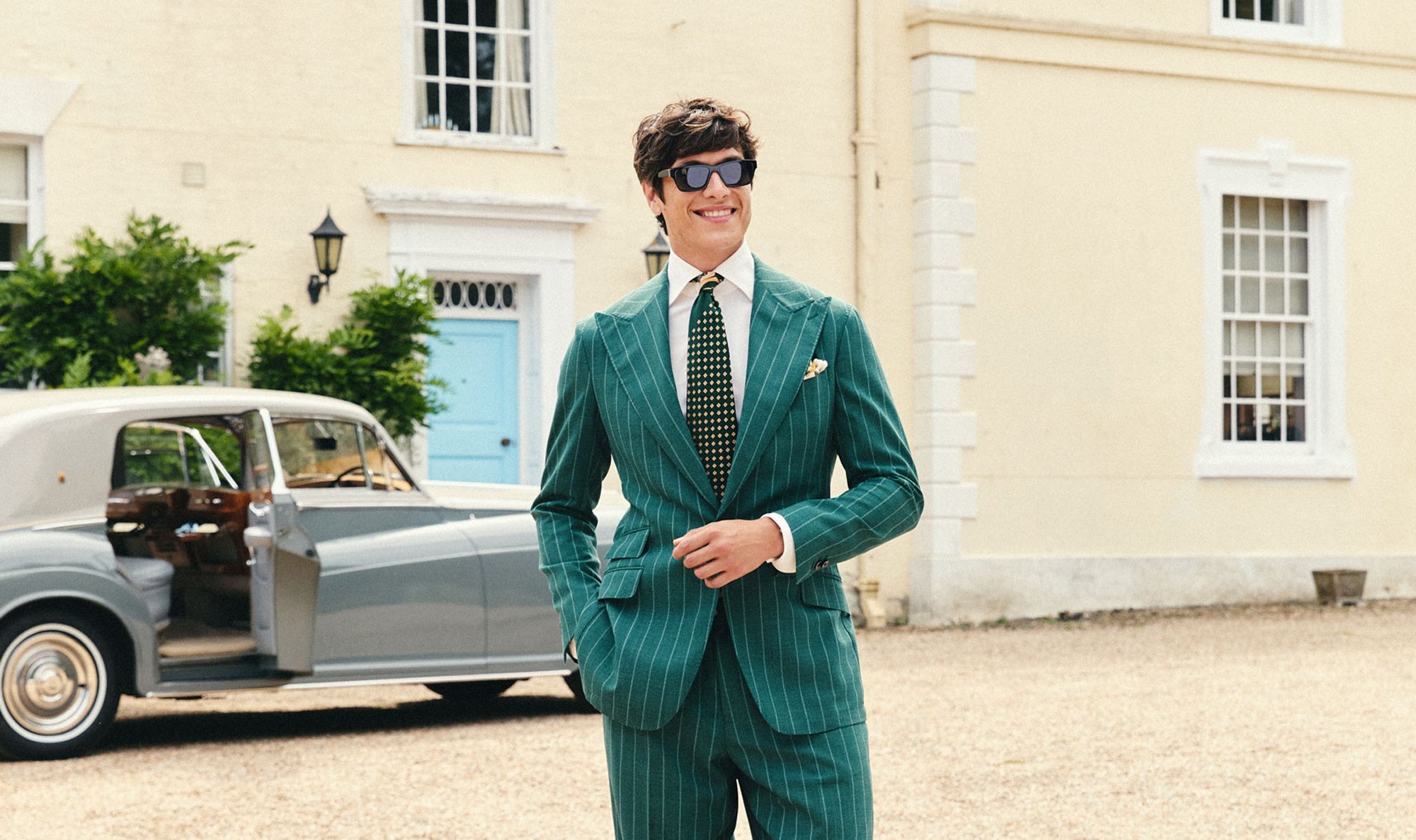 A man in a green pinstripe suit, white shirt, and black tie with yellow dots is smiling and walking outdoors near a vintage car, with a cream-colored building in the background.