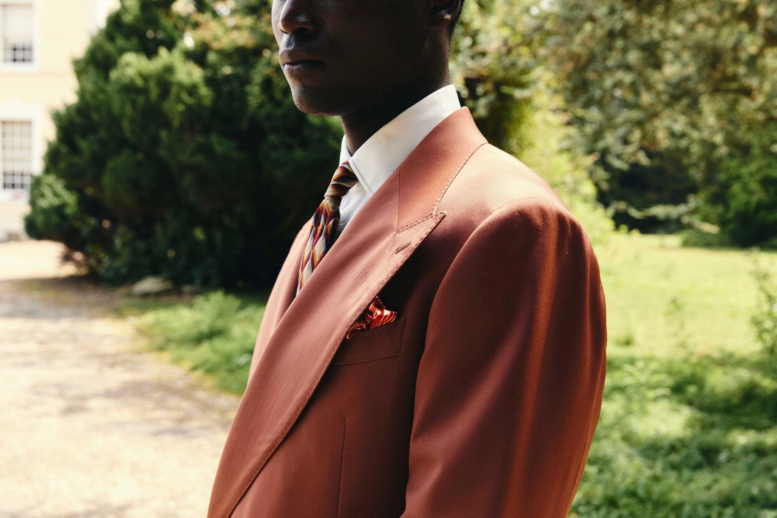 Close-up of a person wearing an orange blazer, white shirt, and colorful striped tie, standing outdoors with greenery in the background.
