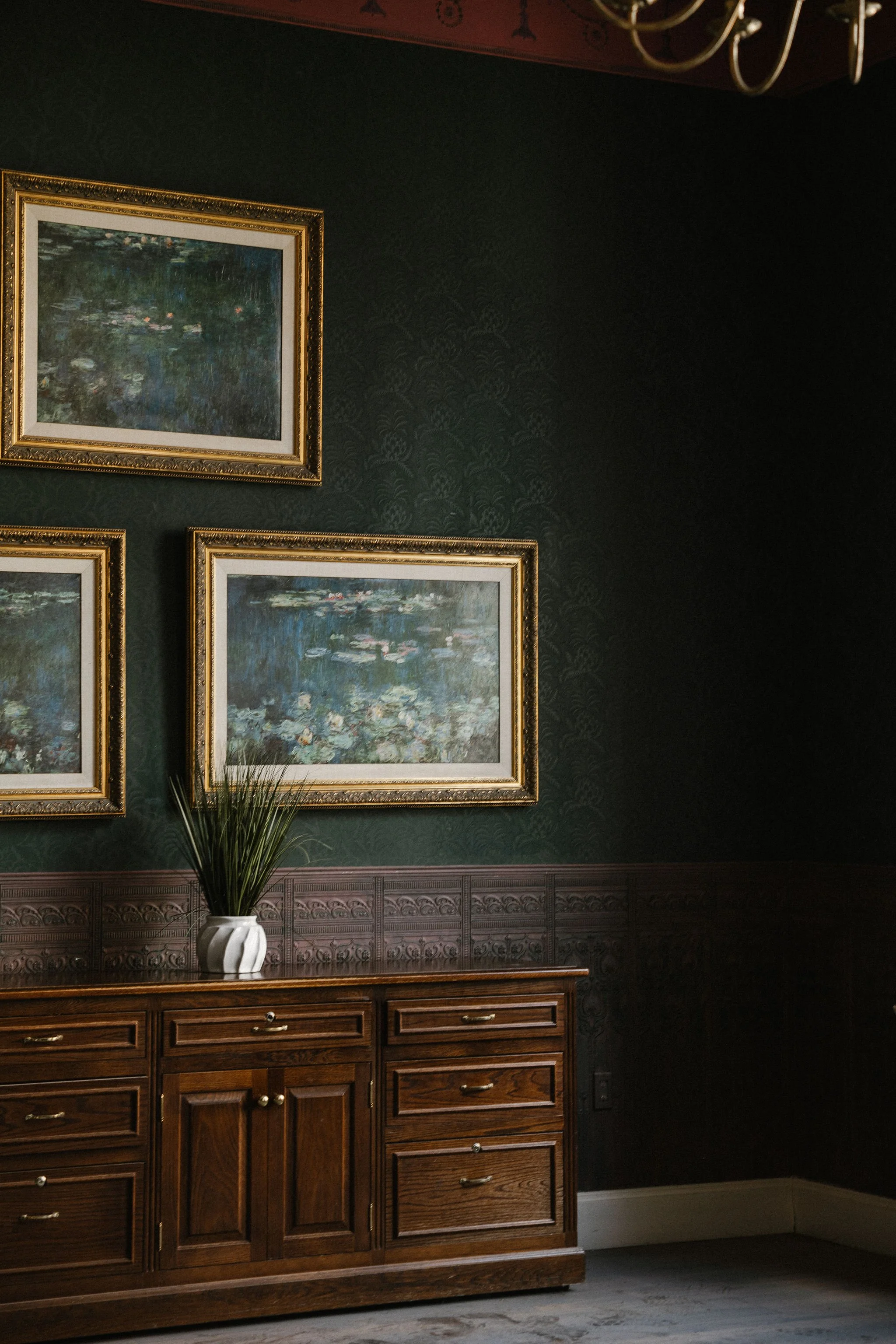 A dark green wall with framed landscape paintings, a wooden sideboard with drawers and cabinets, and a white decorative vase with a plant on top.