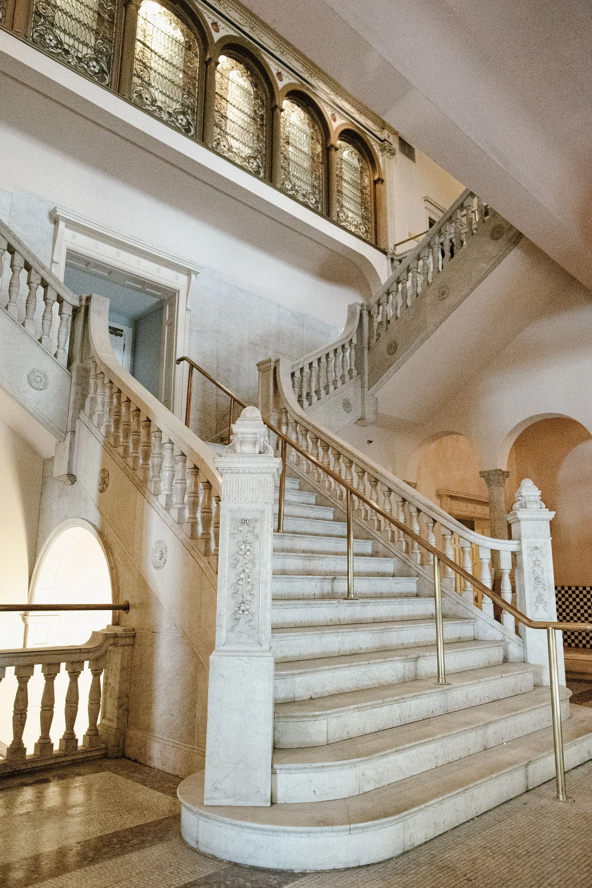 An elegant marble staircase with ornate railings and a decorative balustrade, leading upward inside a historic building with large arched windows with intricate ironwork. The staircase curves gracefully and is illuminated by warm lighting.