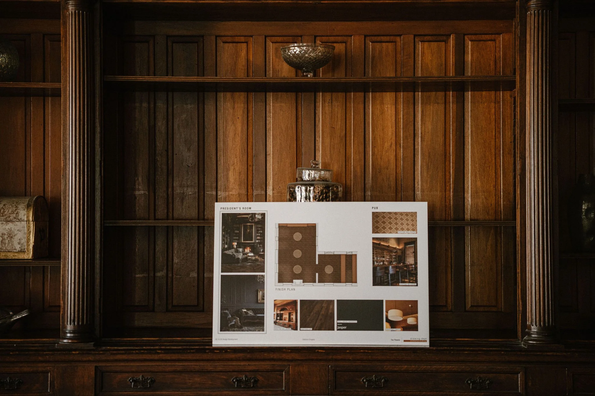 A wooden display cabinet with shelves in the President's room at The Phoenix, there is a presentation board with interior design images, floor plans, and color samples.