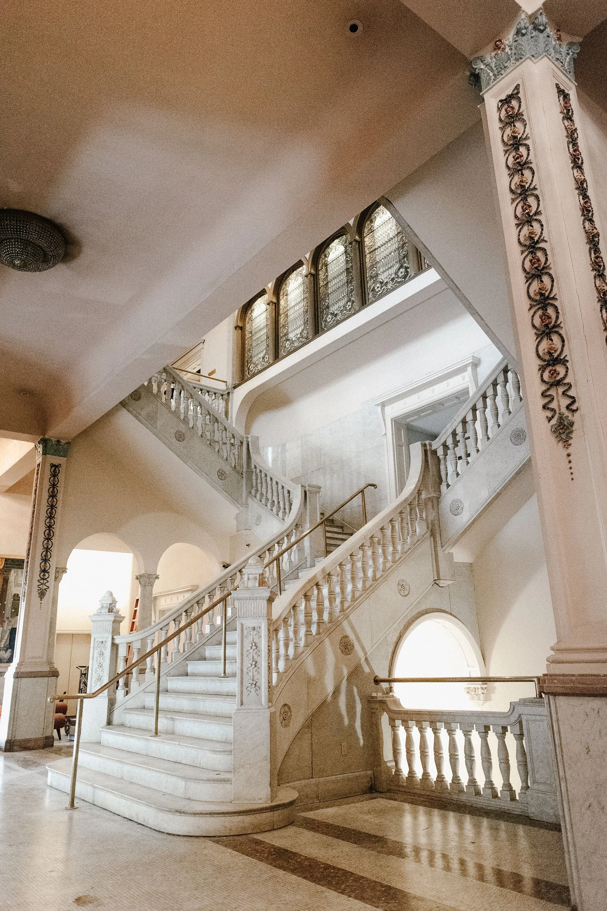 Elegant marble staircase with wooden handrail, ornate white balusters, decorative columns, and stained glass windows in a grand building interior.