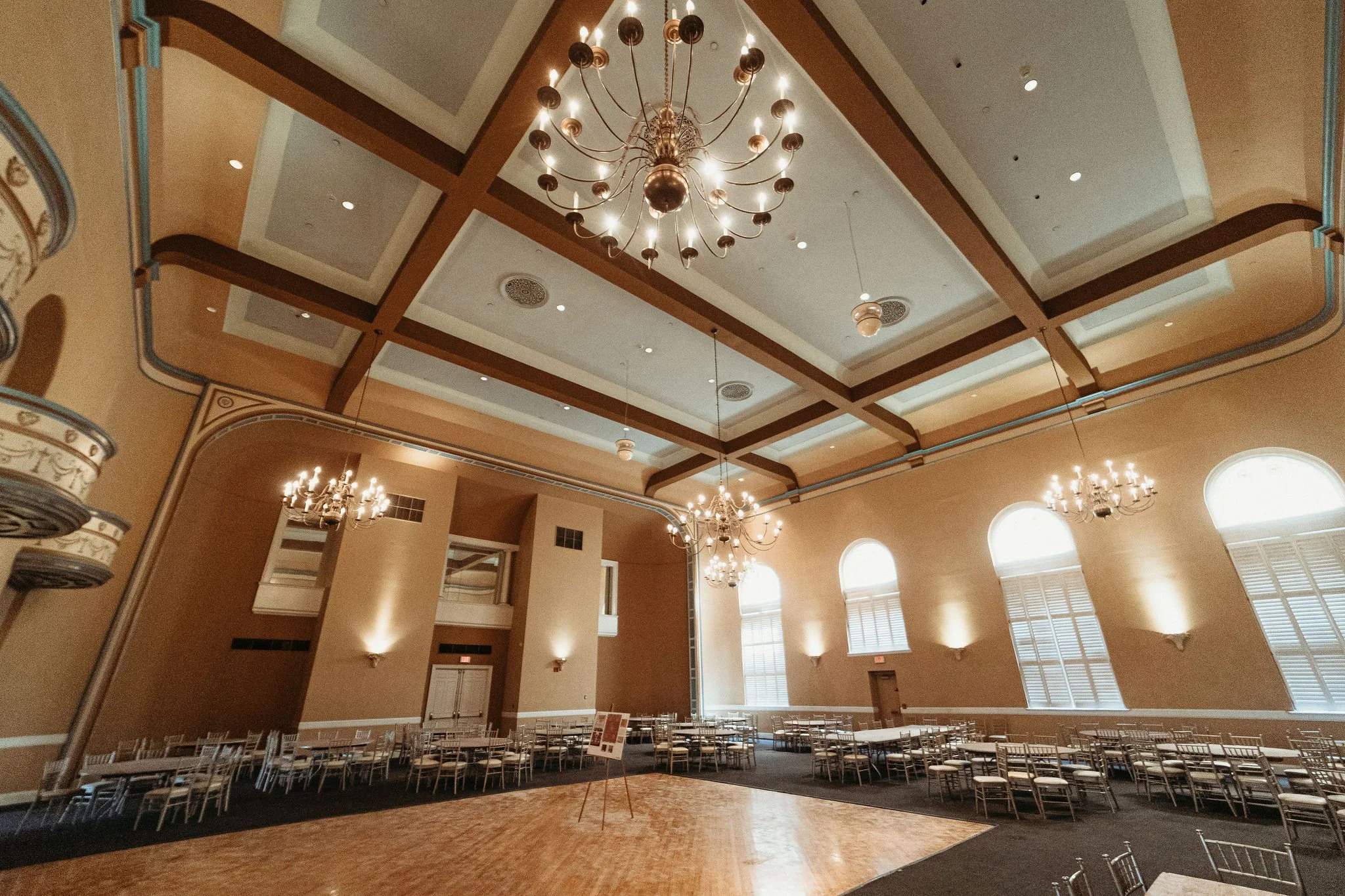 The interior of The Legacy Ballroom, a spacious room with high ceilings, large windows, and elegant chandeliers. The room is set up with rows of chairs and has decorative wall moldings and lighting fixtures.