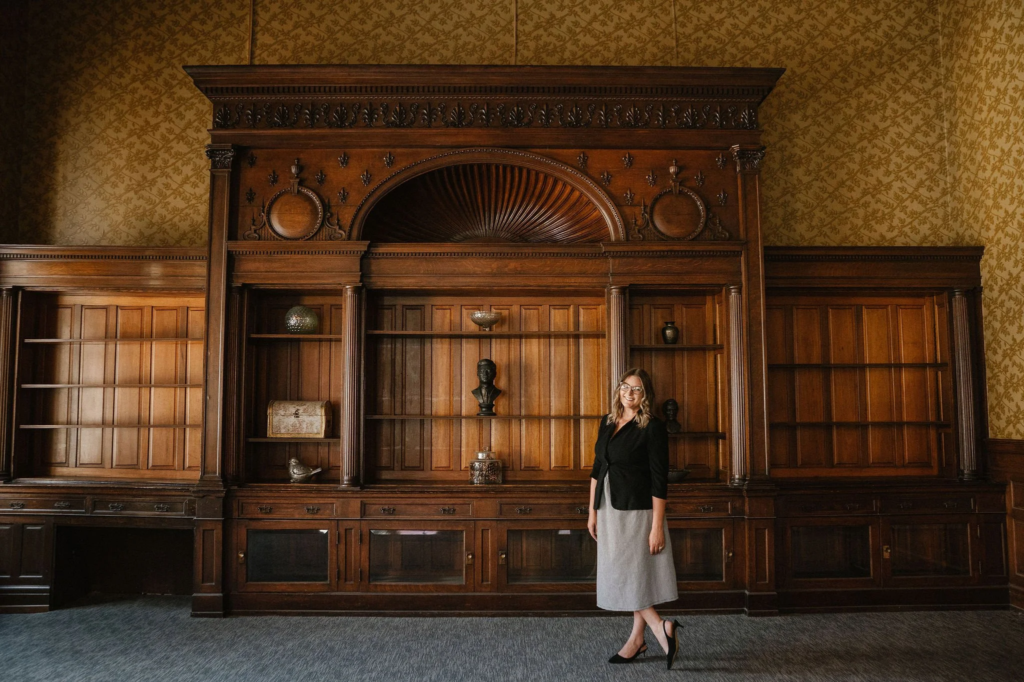 A woman standing in front of a large, ornate wooden bookshelf or paneling in a historic or formal interior, wearing a black blazer, a gray skirt, and high heels, smiling at the camera.