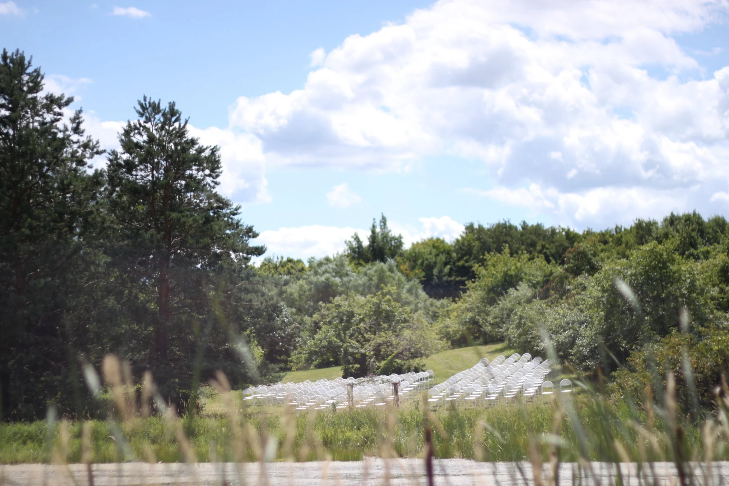 A landscape with a blue sky, scattered white clouds, tall green trees, and an open field with rows of white chairs, possibly set up for an outdoor event or gathering.
