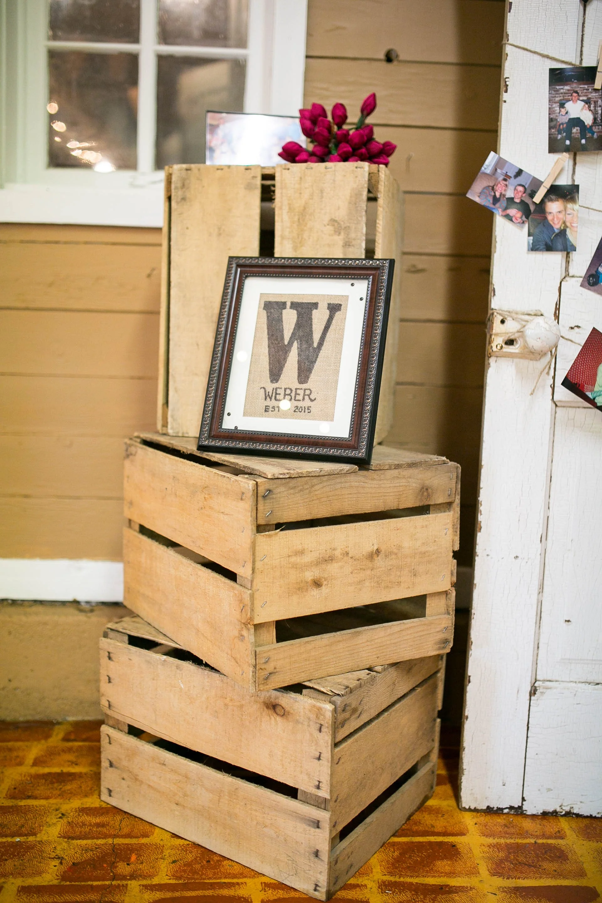 Stack of wooden crates with framed picture of the letter W and text 'Weber EST 2015' on top. Behind the crates, a bunch of red tulips in a wooden box. To the right, photos clipped to a white wooden wall. There is a window with white trim and a brick wall visible outside.