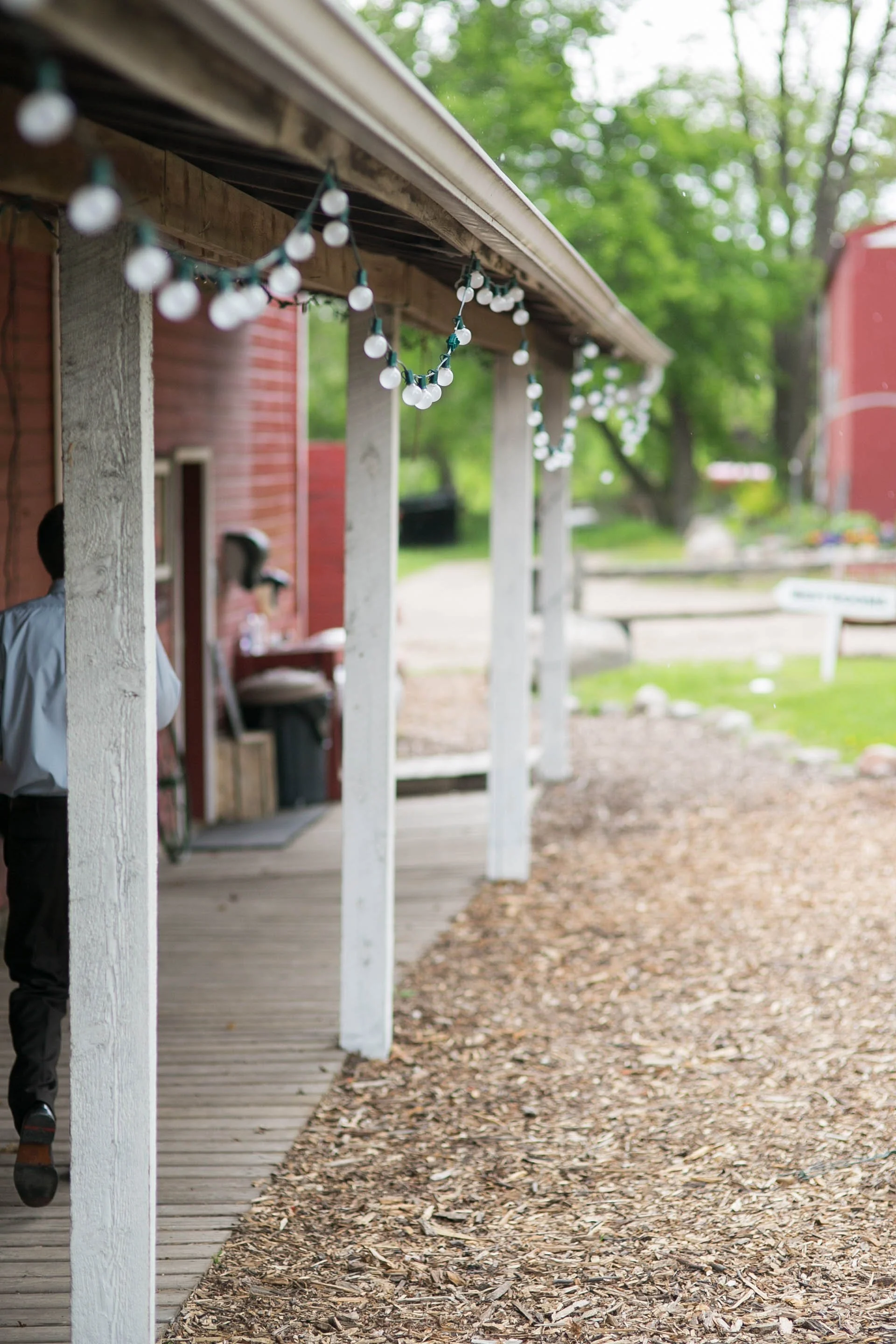 A porch with white wooden columns and string lights hanging, a person in uniform partially visible, a brick building to the left, trees and greenery in the background, dirt and wood chip ground covering.
