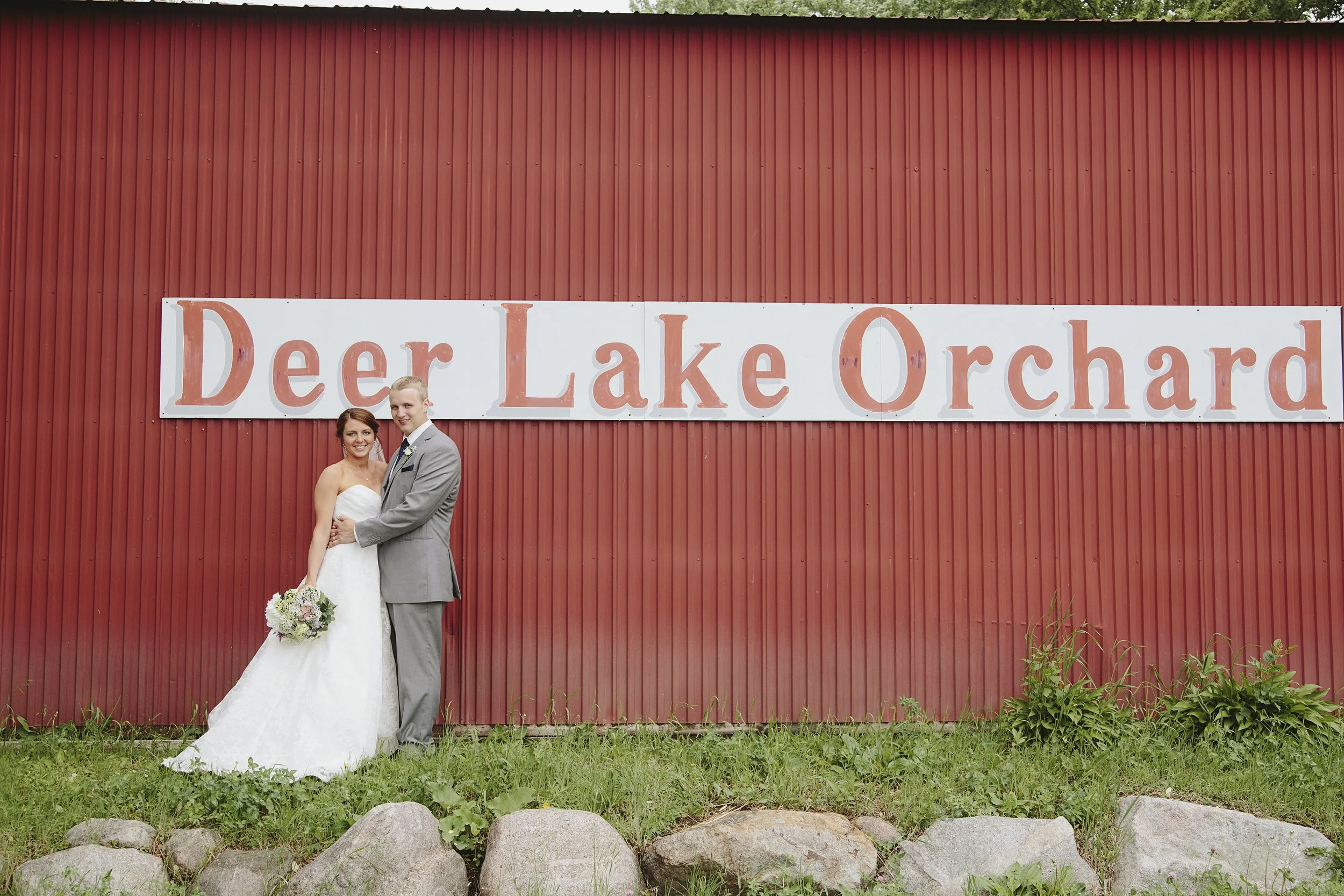 A bride and groom in wedding attire standing in front of a large sign reading 'Deer Lake Orchard' on a red barn wall. The bride is holding a bouquet and the couple is smiling at the camera.