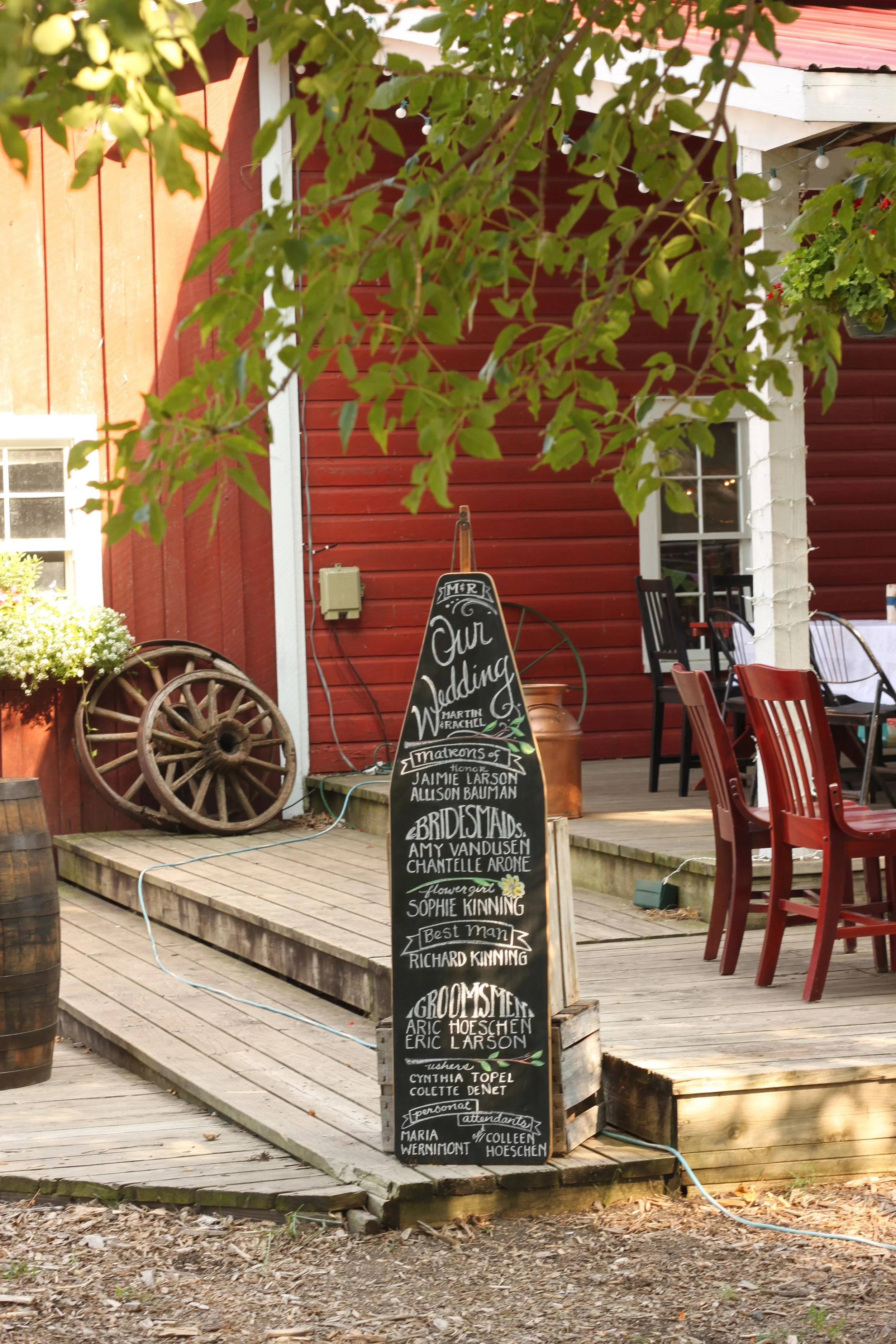 A wooden deck with a blackboard sign listing names of people at a wedding, surrounded by chairs and outdoor decor, with a red building and greenery in the background.