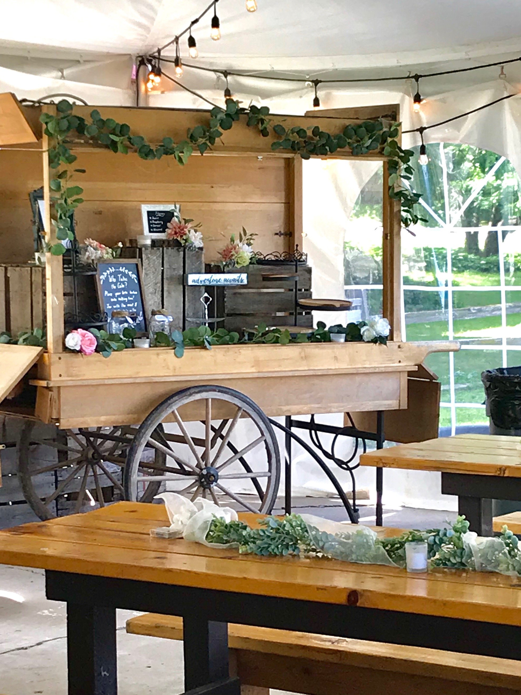 A flower cart decorated with greenery and flowers inside a tent with string lights, set up for an event.