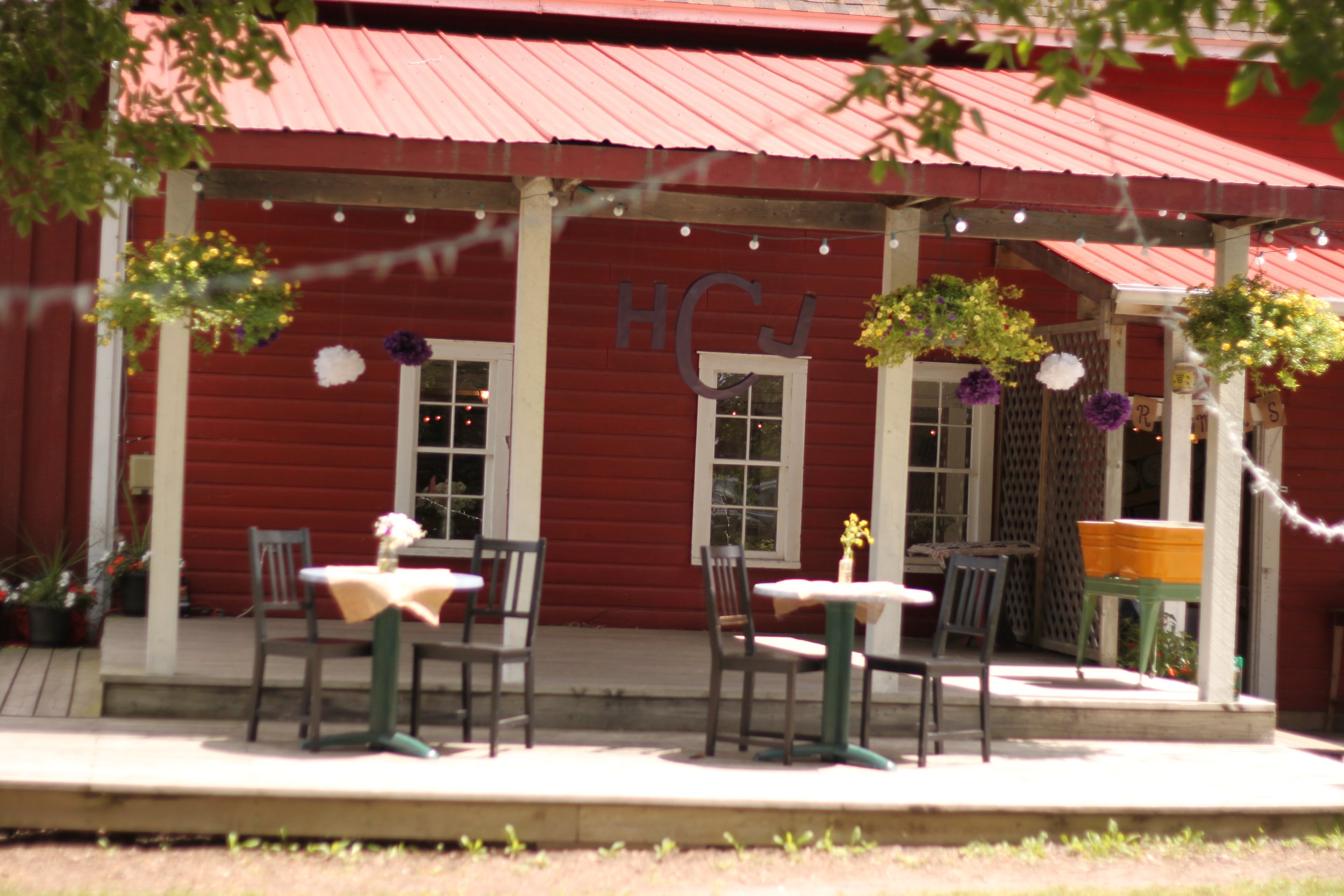 An outdoor patio area with several tables and chairs, decorated with hanging flower baskets and string lights, in front of a red wooden building with white window frames.