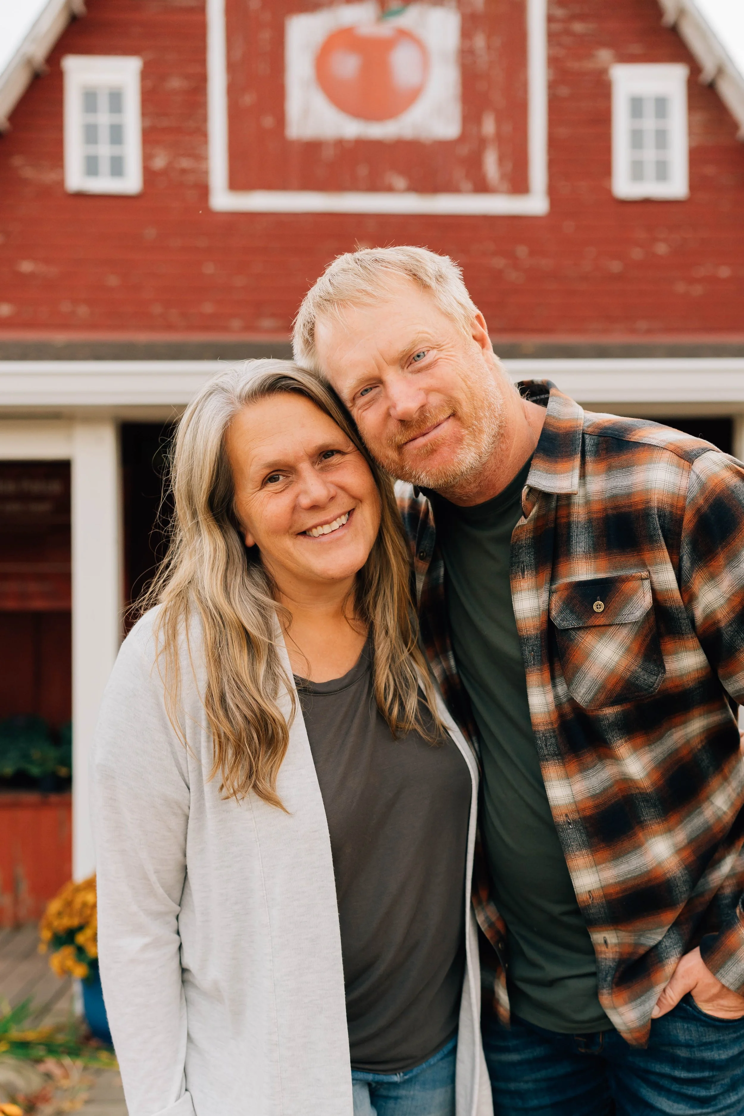 A smiling woman with long blonde hair and a man with short blonde hair and beard, both in casual clothing, standing close together in front of a red barn with a painted pumpkin on it, during daytime.