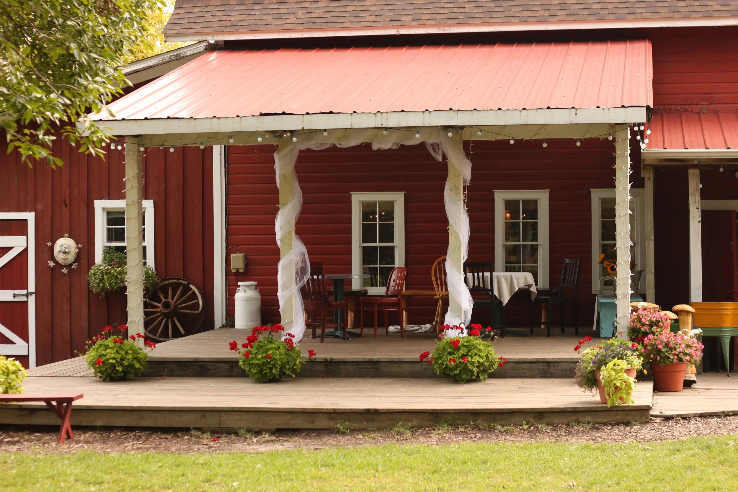 Red barn with a wooden deck and patio furniture, potted plants, string lights, and decorative items outside.