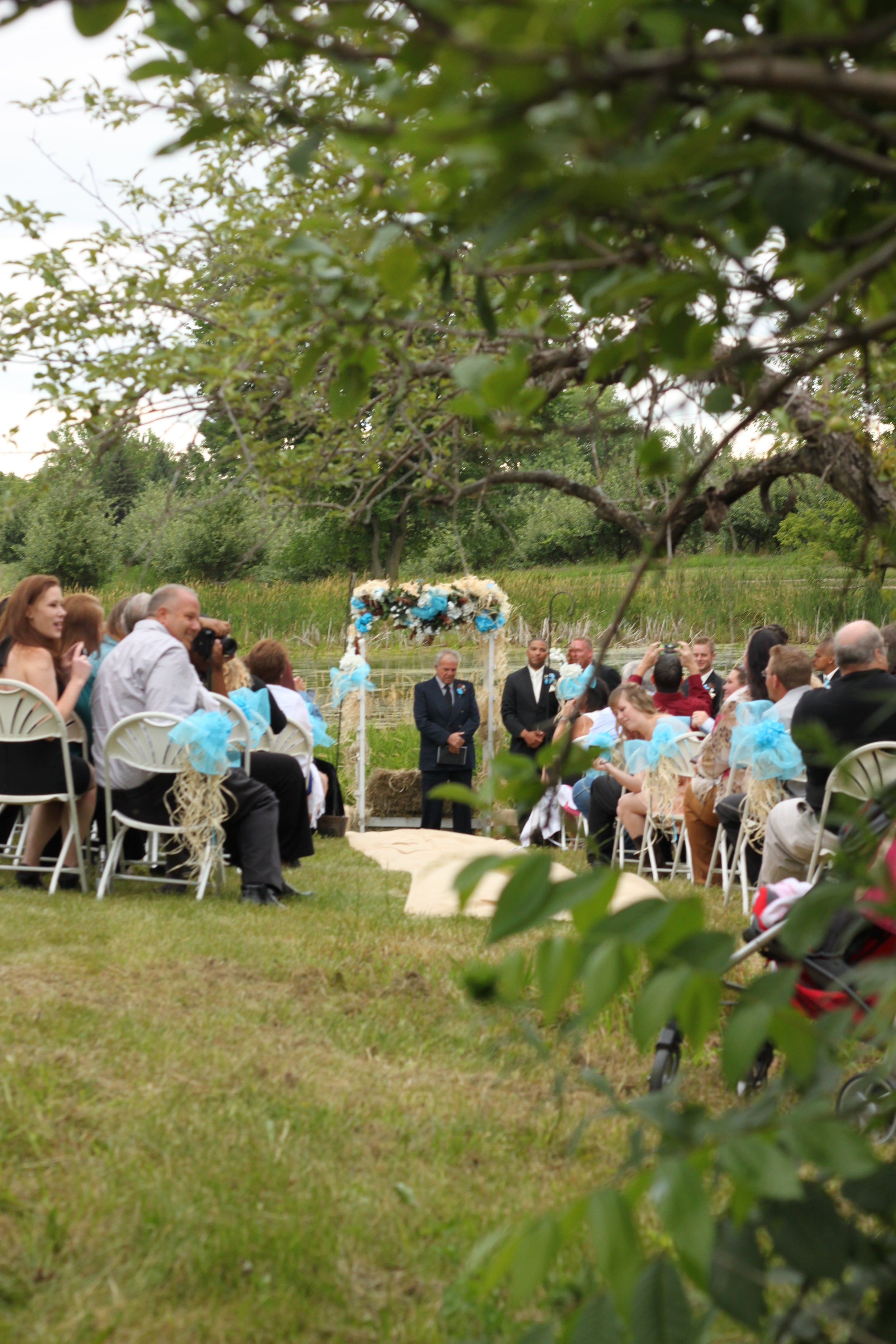 An outdoor wedding ceremony with guests seated on white chairs, decorated with blue bows, facing a couple standing under a floral arch in front of a pond, surrounded by trees and greenery.