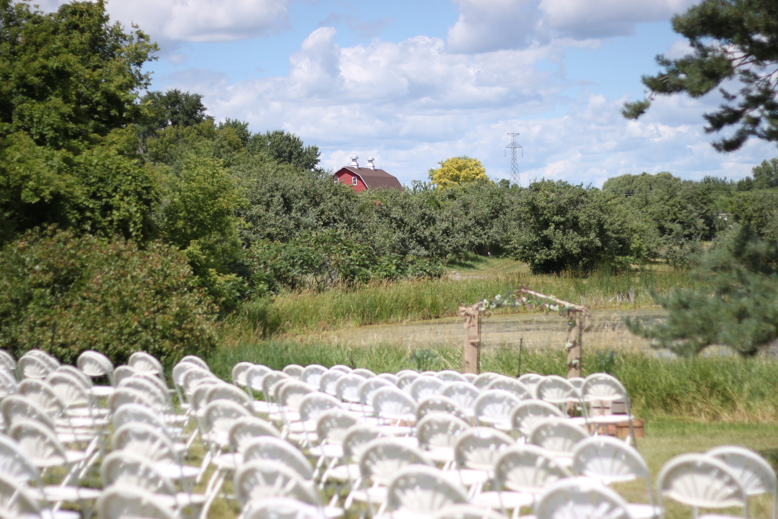 Rows of white chairs arranged outdoors in front of a lush green area with trees, a red barn, and a blue sky with clouds.
