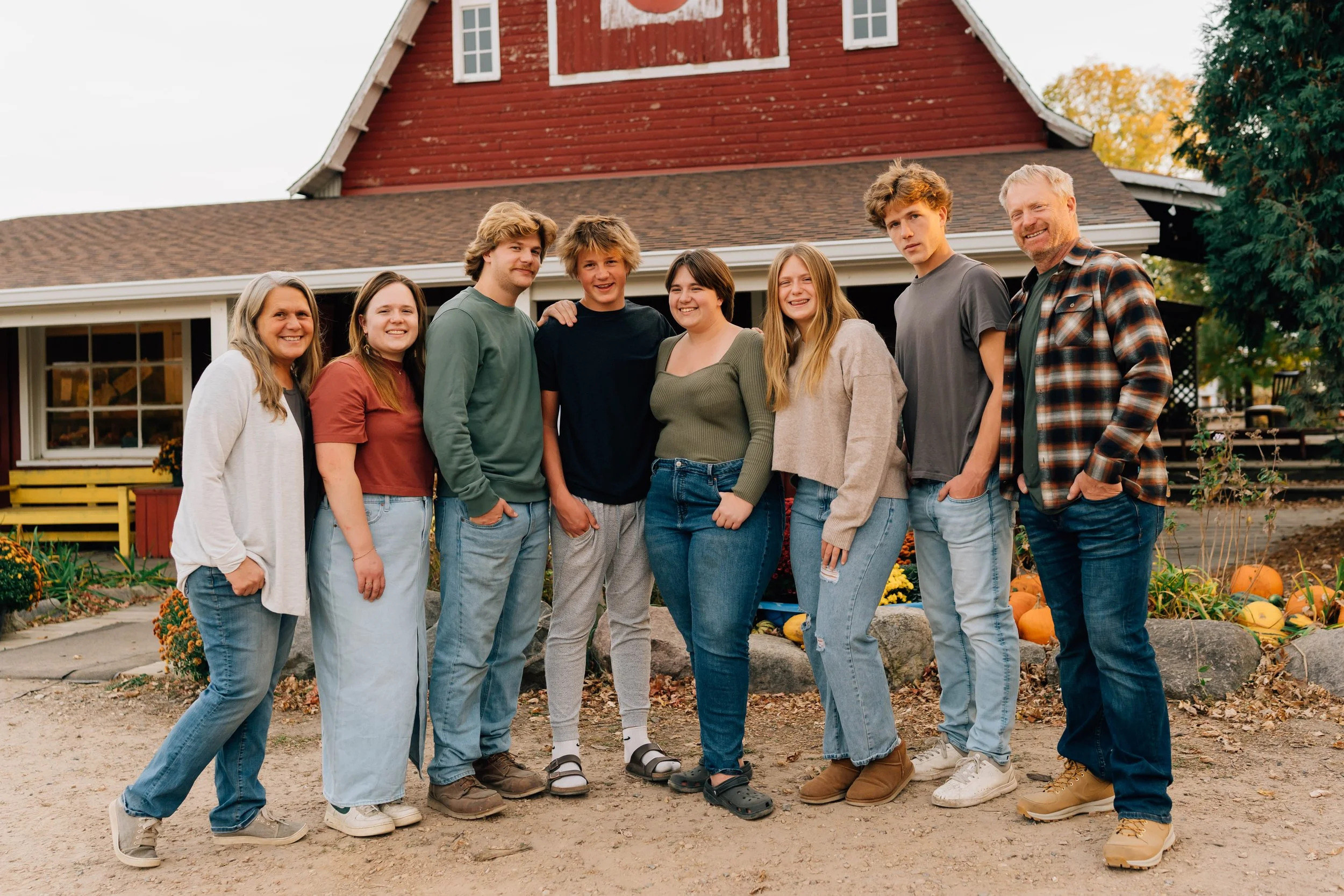 Group of eight people smiling and standing closely together outside in front of a house with autumn decorations and pumpkins.