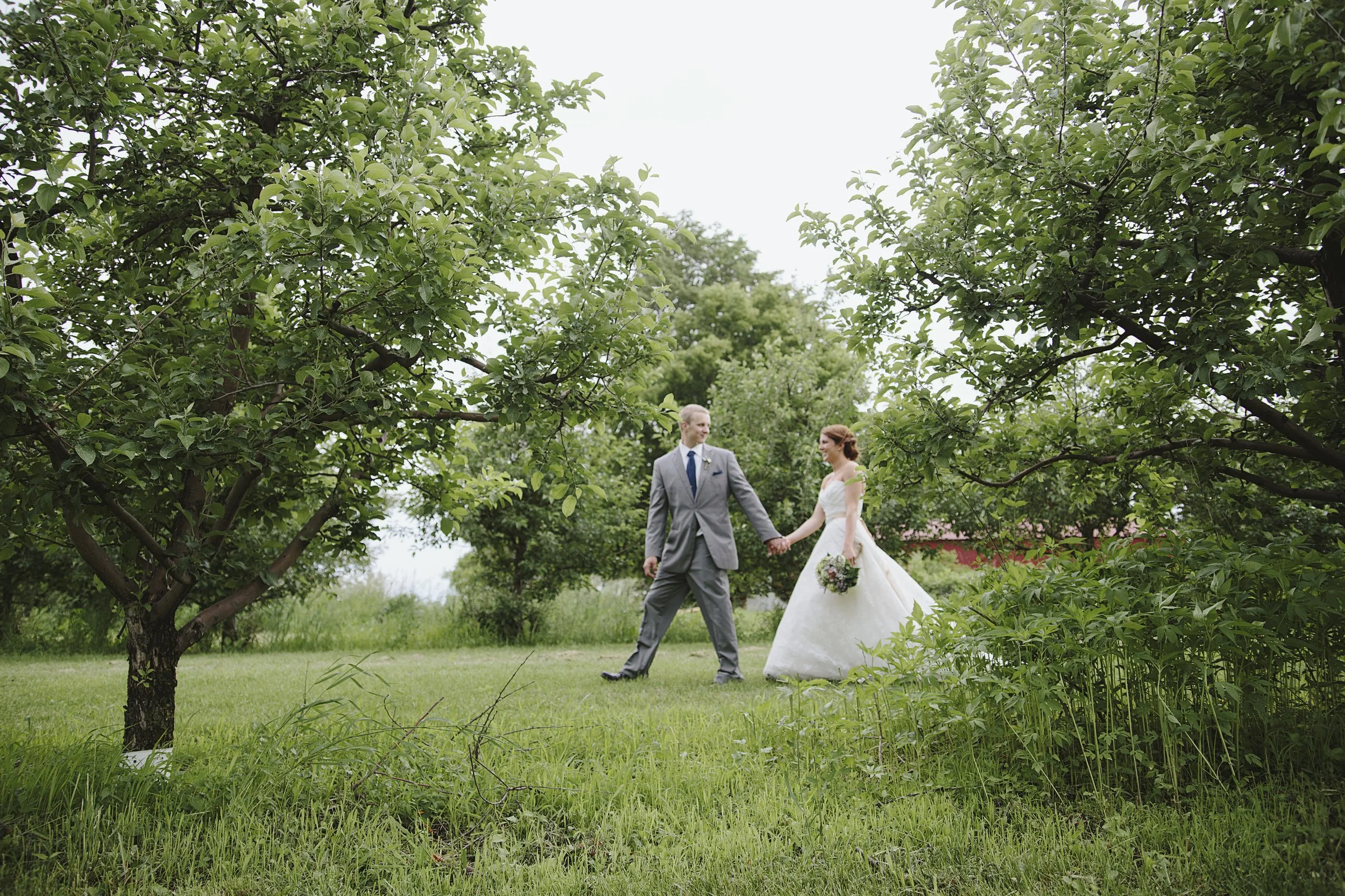 A bride and groom holding hands and walking in a grassy outdoor setting, surrounded by lush green trees and plants, during daytime.