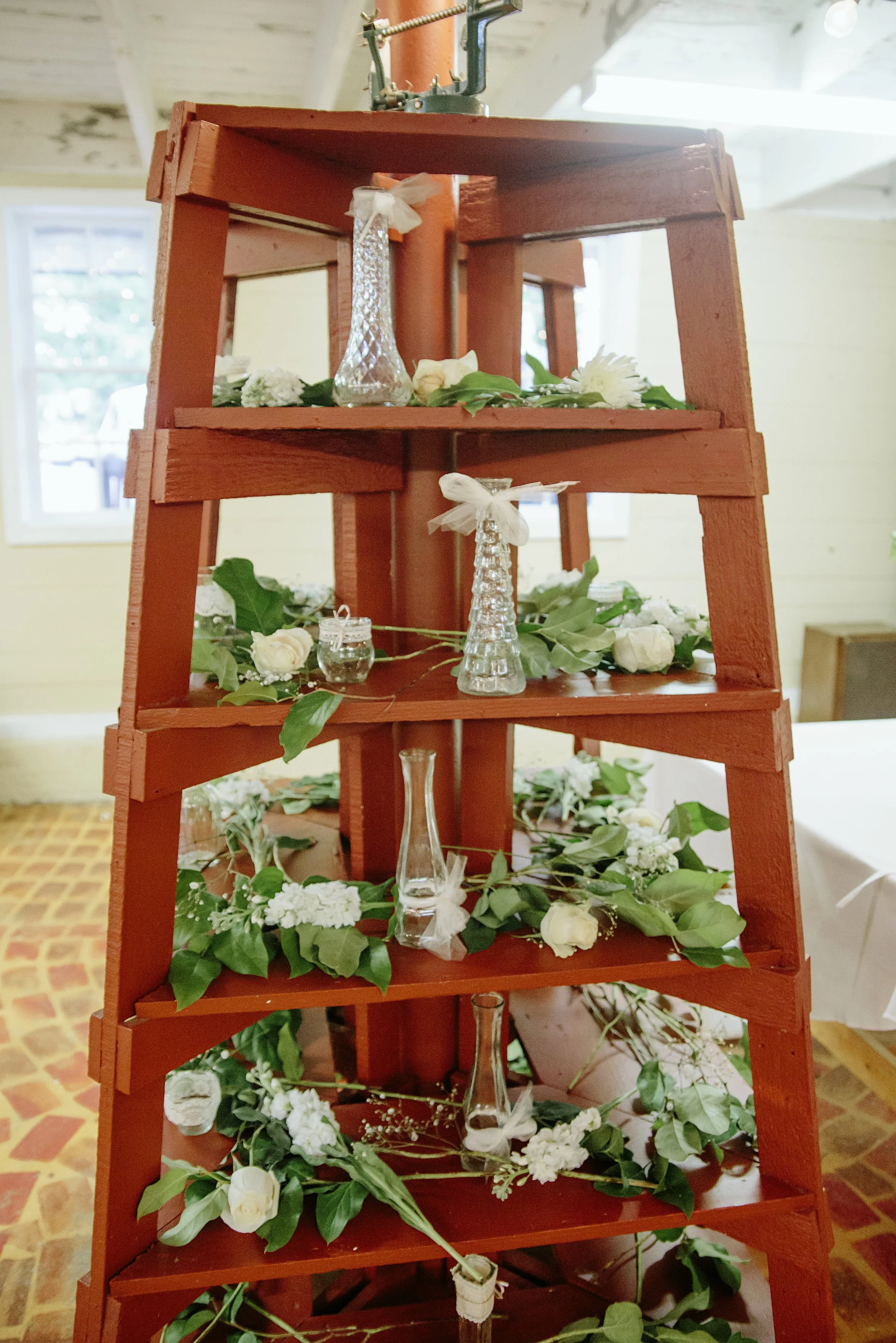 A red wooden shelf decorated with glass vases, white flowers, and green leaves. Small candles are also placed on the shelves.