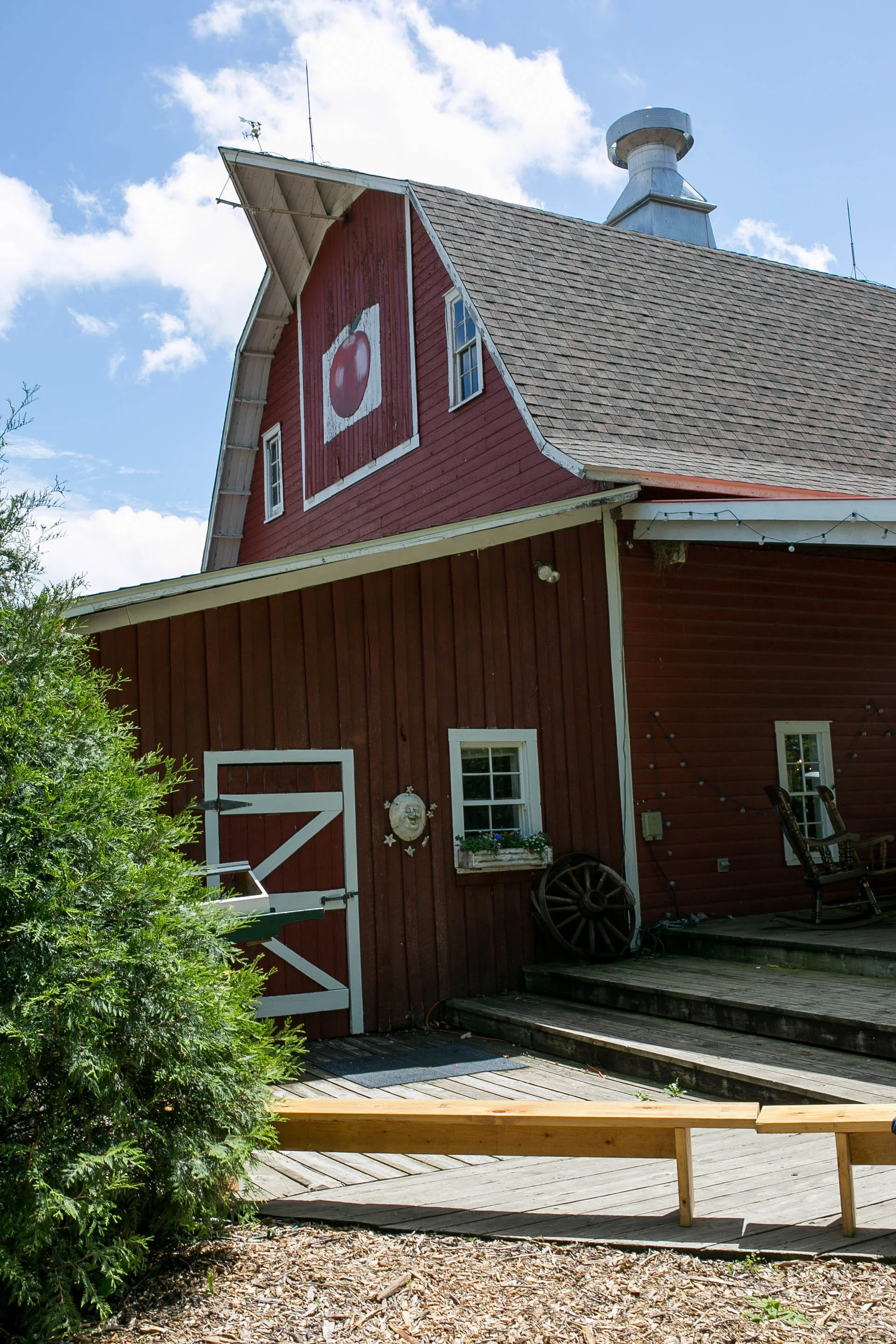 Red barn with a large white apple logo painted on the upper side, a small window, a white door with a latch, outdoor rocking chairs, an old wagon wheel, and a window box with flowers, set against a blue sky with clouds.