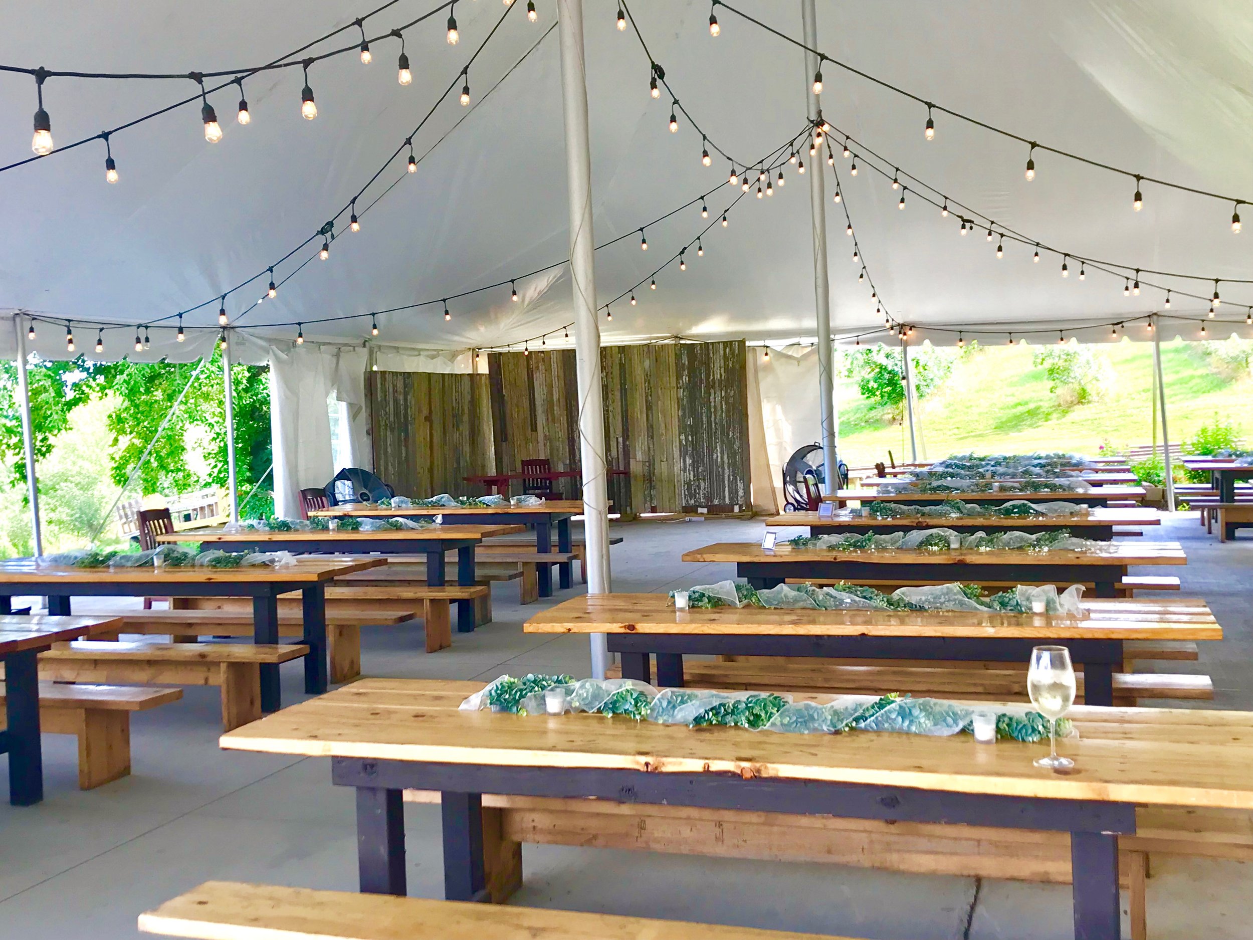 A large event tent with string lights hanging from the ceiling, set up outdoors with green trees visible in the background. Inside, wooden tables are decorated with greenery and small candles, arranged for a gathering or celebration.