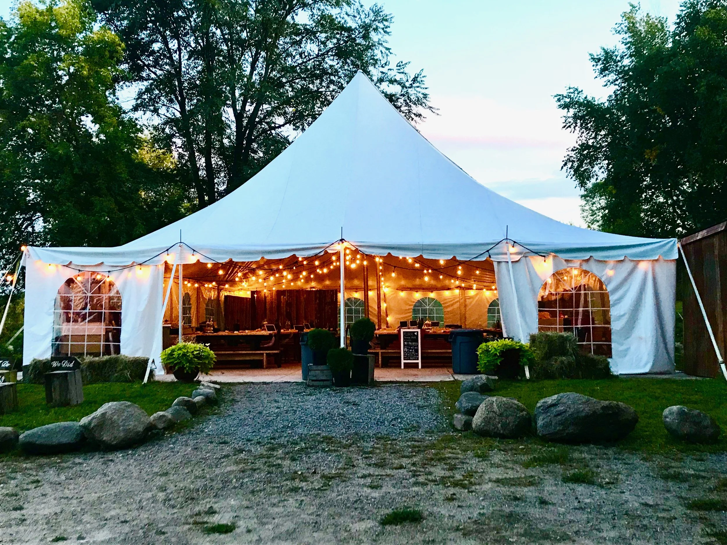 A large white event tent with string lights hanging inside and outside, set up outdoors among green trees during dusk or evening.