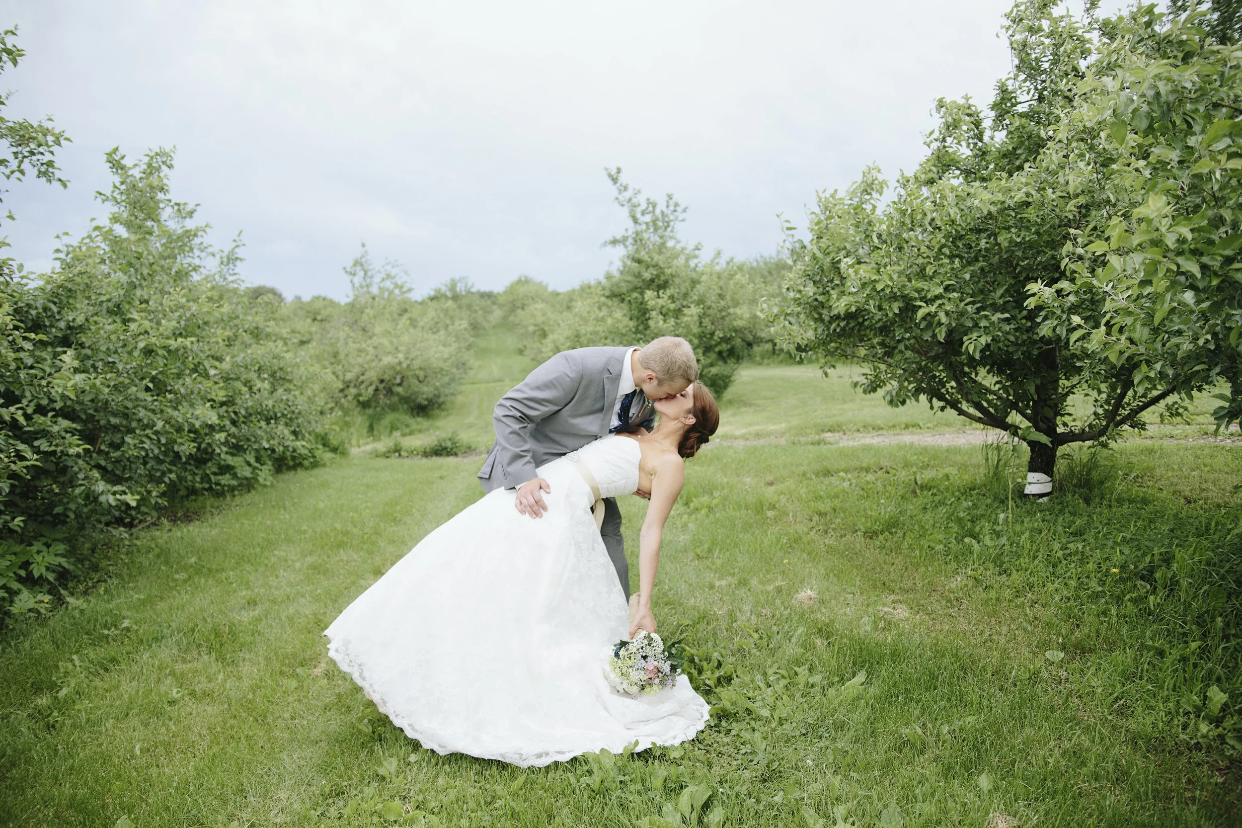 A newlywed couple sharing a kiss outdoors during daytime, with the groom dipping the bride in a grassy area surrounded by trees.