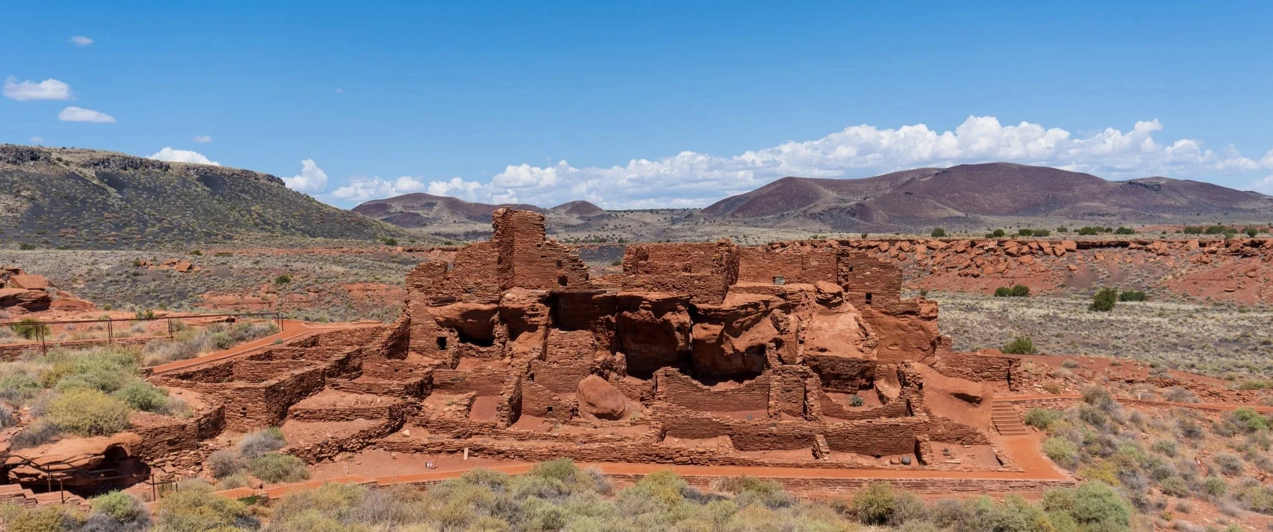 Ancient red rock pueblo remains in a desert landscape with hills and blue sky in the background.