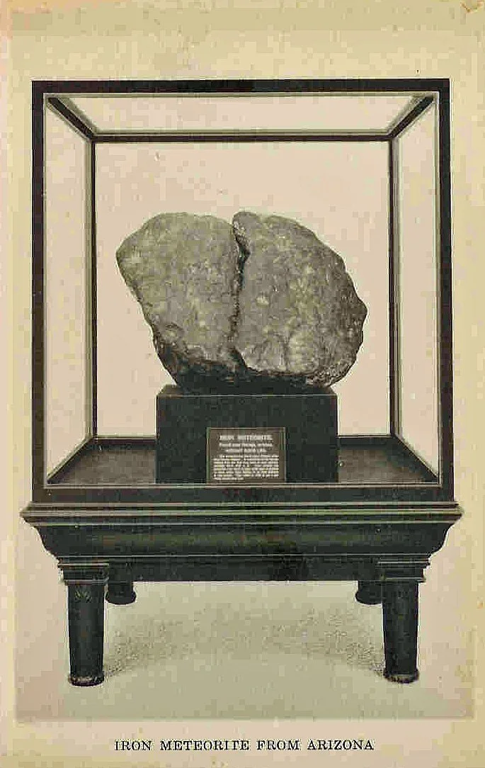 The Navajo Meteorite on display at the Smithsonian in the 1930s in a large display case on a black wooden stand, with a small descriptive plaque. The meteorite is irregularly shaped. The label reads "Iron Meteorite from Arizona."