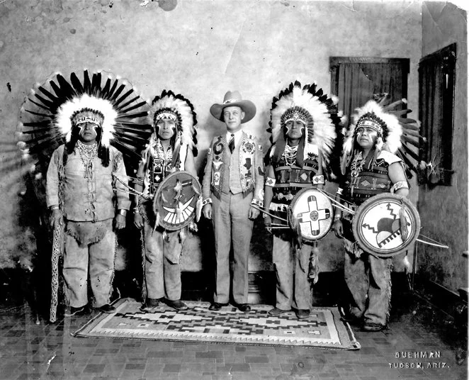 Billingsley Hopi Dancers in feather headdresses and clothing with Shields and Spears, standing indoors on a patterned rug, with Milo Billingsley in Western attire and a cowboy hat in the center. 