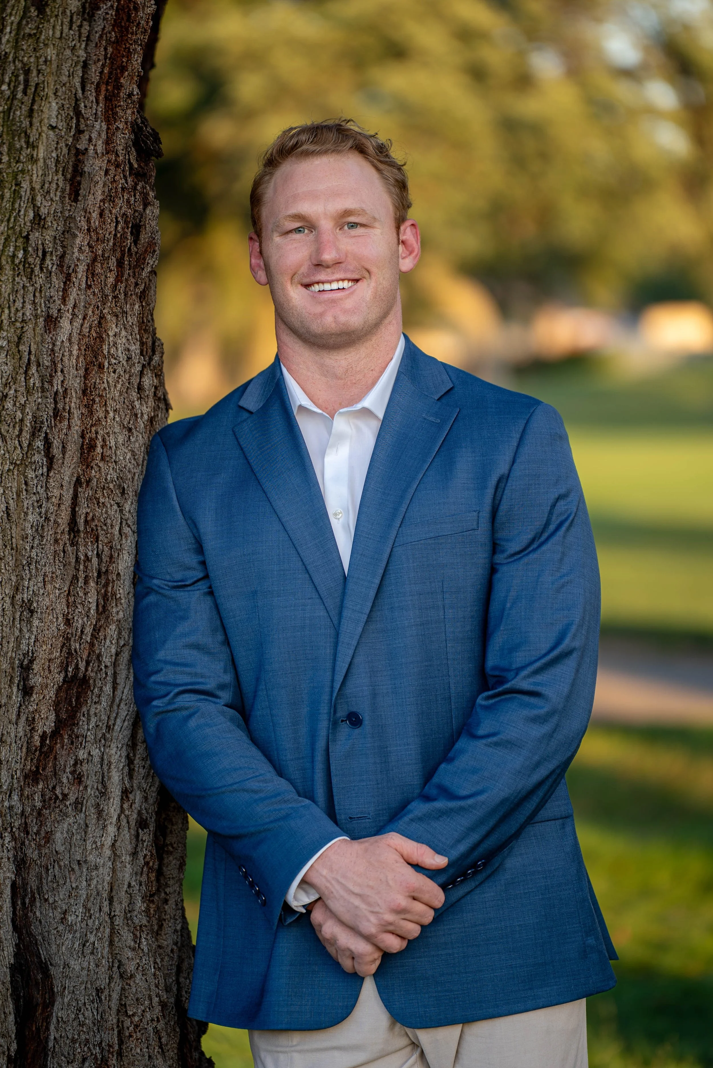 A man in a blue suit jacket and white dress shirt standing outdoors near a tree on a sunny day, smiling with arms crossed in front of a blurred background of trees and grass.