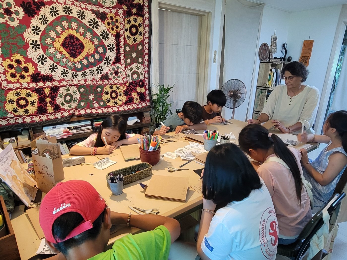 Children and an adult seated around a table engaged in creating with art supplies, in a room decorated with a colorful textile wall hanging and bookshelf.