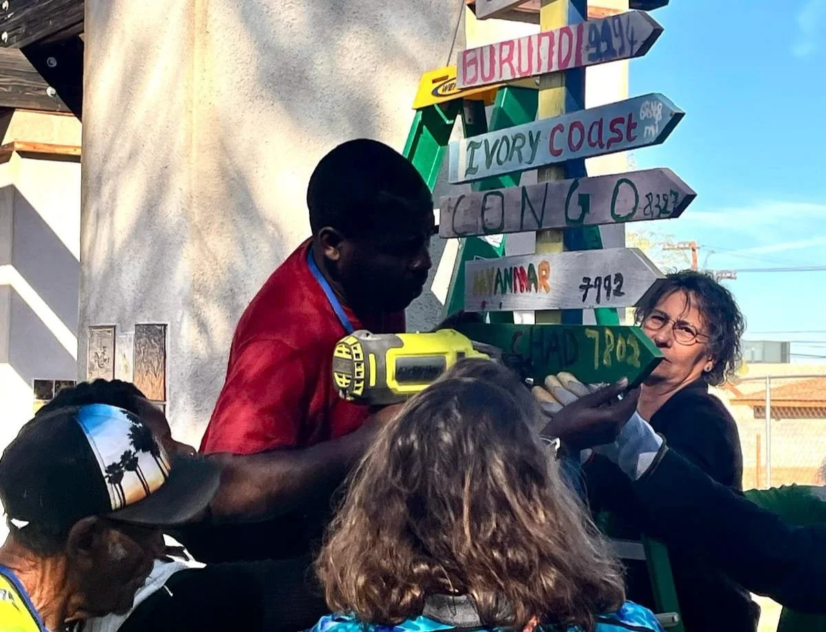 A group of people, including a man in a red shirt using a cordless drill, a woman with glasses, and children, are gathered around a colorful signpost with multiple destination signs pointing in different directions, such as 'IVORY COAST,' and 'MYANMAR.' The scene takes place outdoors with a bright blue sky and some buildings in the background.
