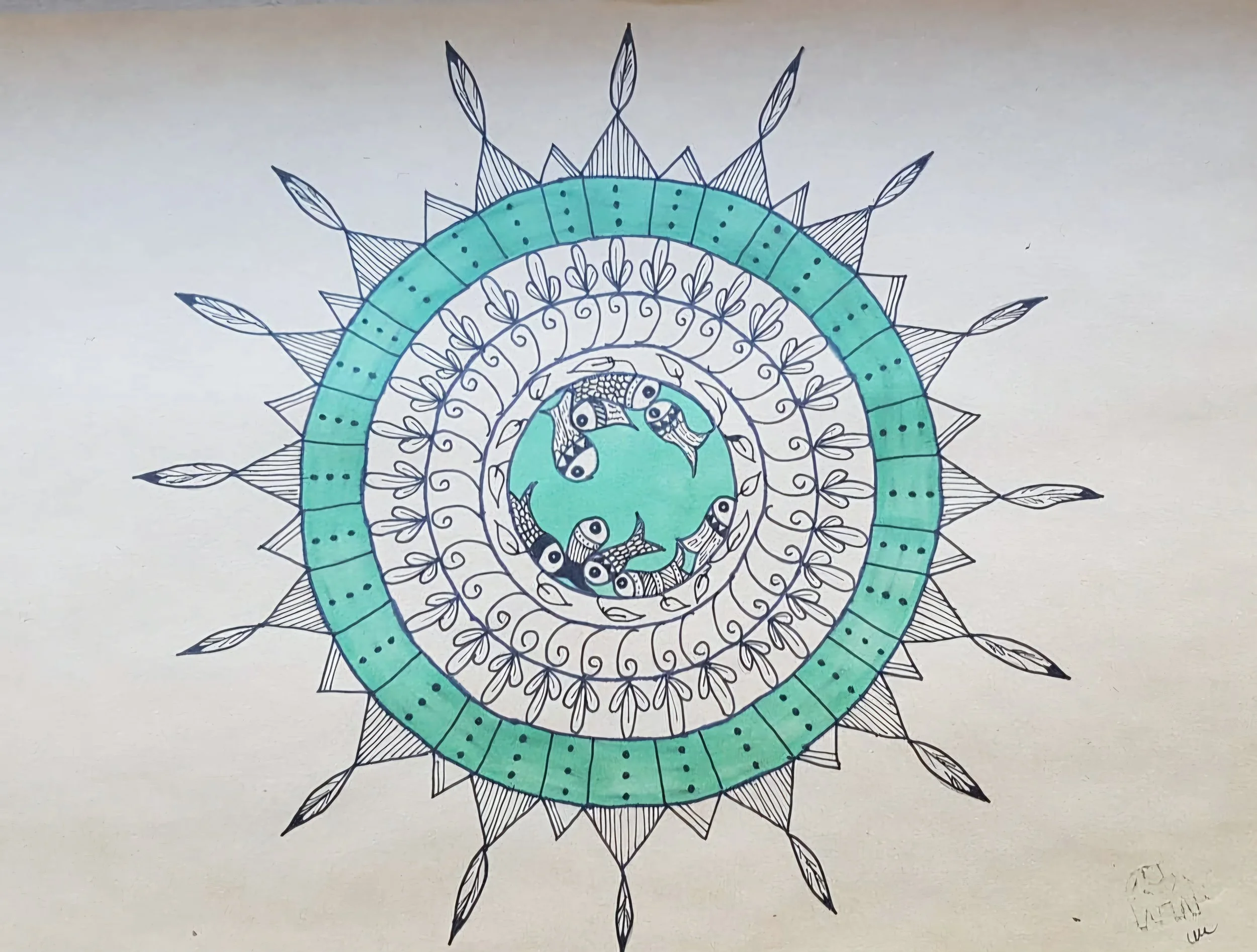 A detailed, colorful mandala drawing on paper, featuring concentric circles with intricate leaf and geometric patterns, and small fish illustration at the center.