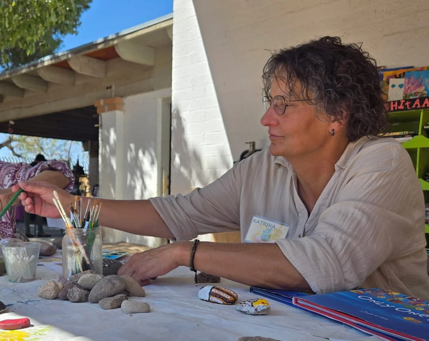 A woman with curly hair and glasses, wearing a beige shirt with a name tag that says 'Katrina', is sitting at a table outdoors painting. She is surrounded by art supplies, books, and rocks, with trees and a building in the background.