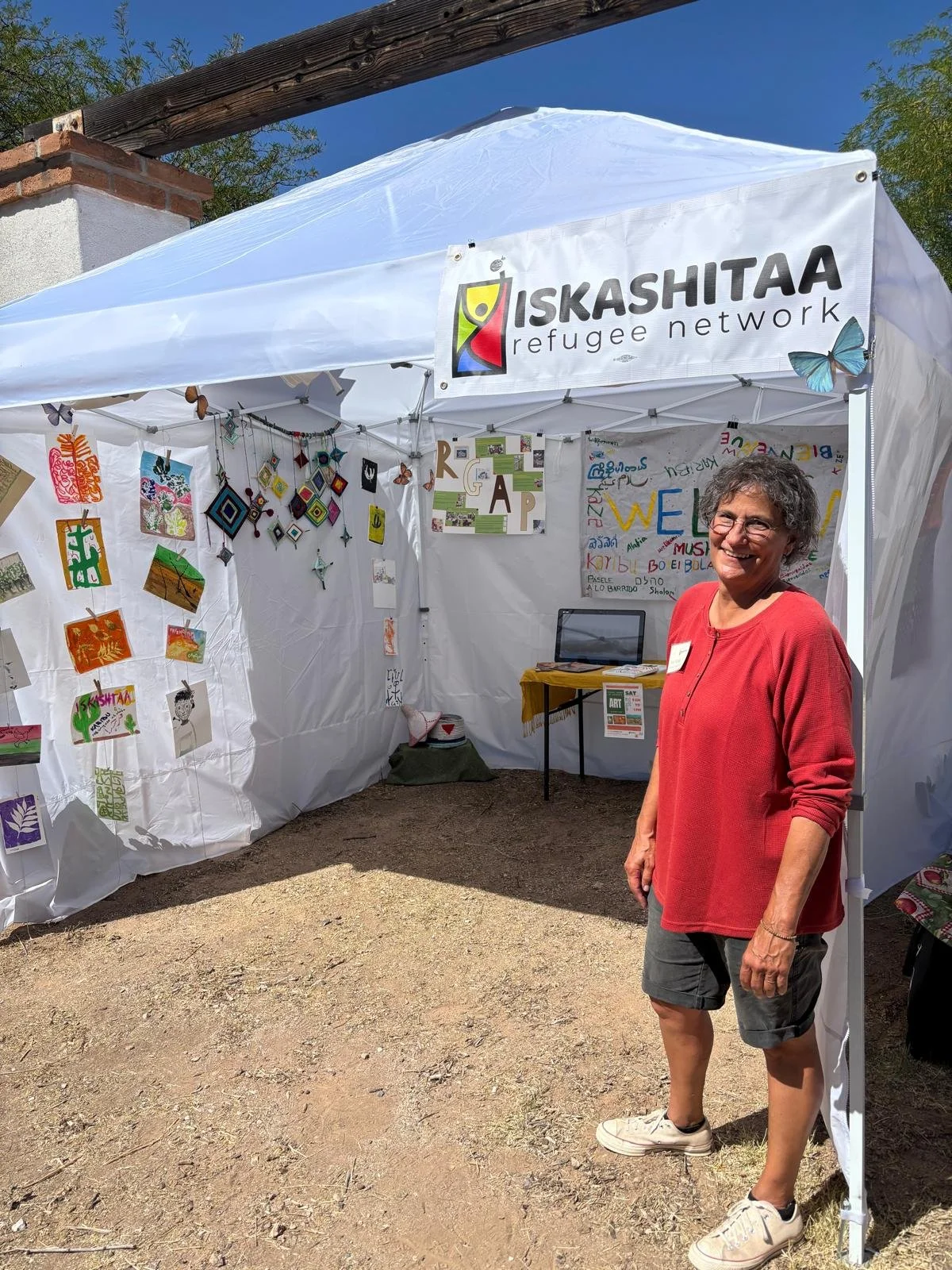 A woman standing outside a white tent with a sign reading "ISKASHITAA Refugee Network". The tent displays colorful artwork and crafts. The woman is smiling, wearing a red top, shorts, glasses, and white shoes.