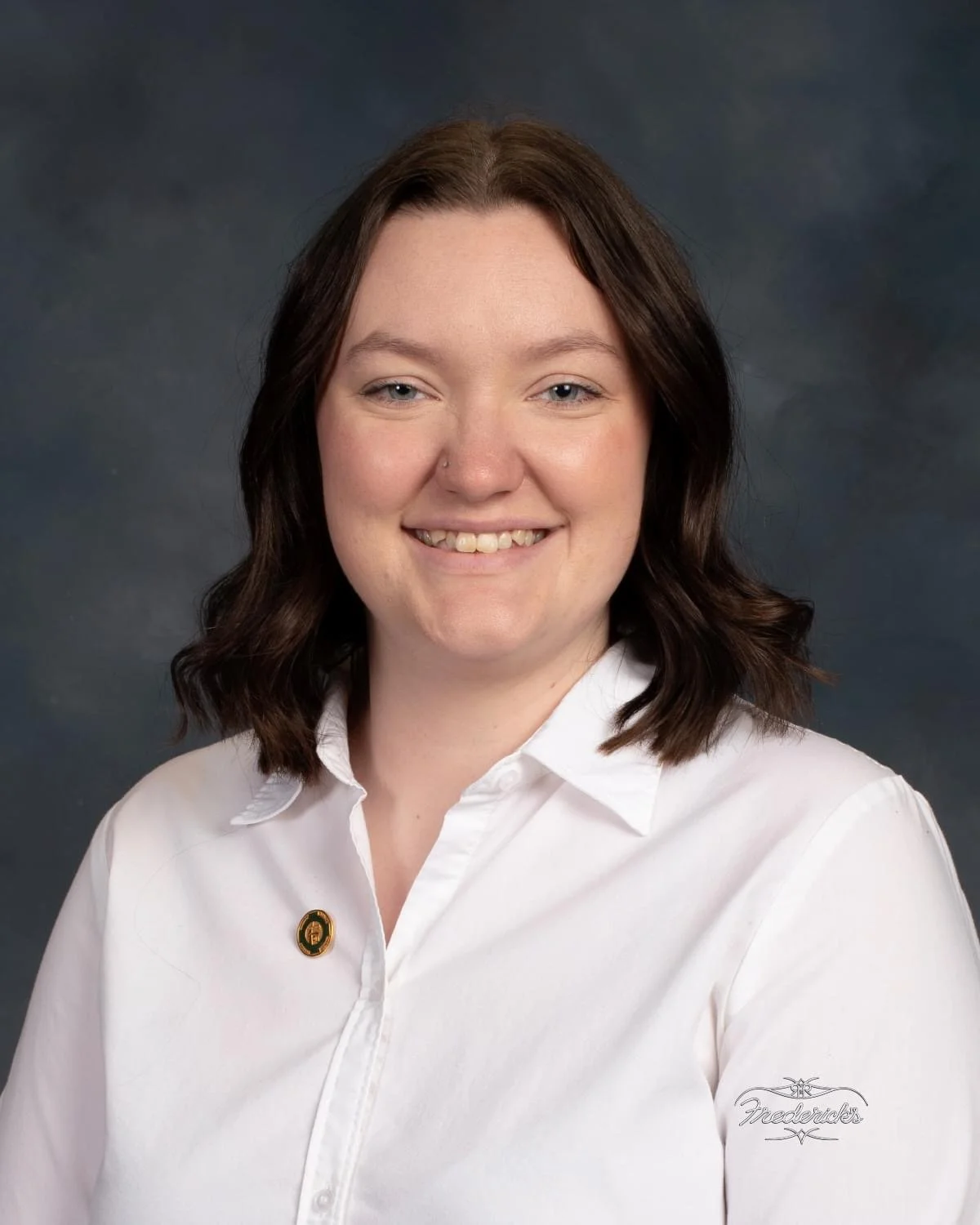 Portrait of a woman with shoulder-length brown hair, wearing a white collared shirt with a gold pin on the left side, smiling against a dark, cloudy background.
