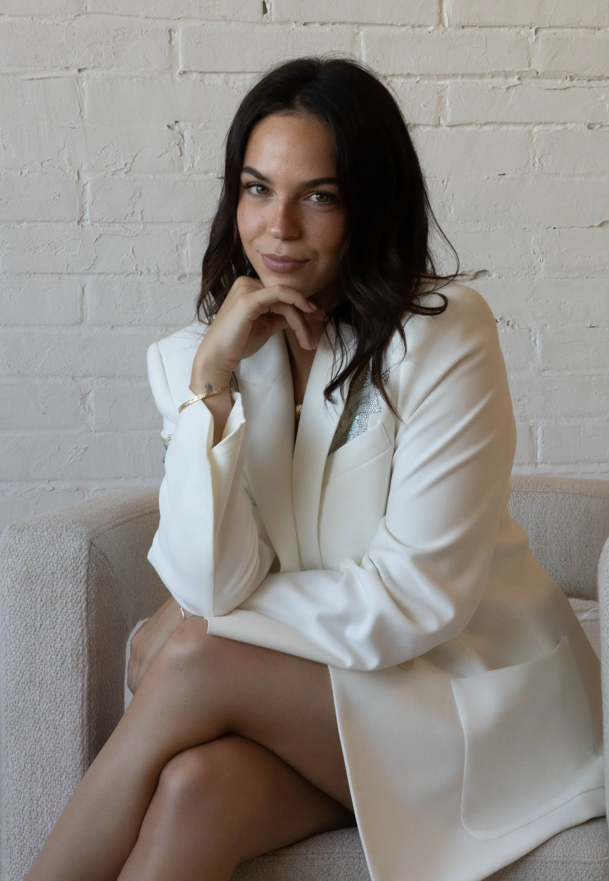 A woman with dark hair and light skin, wearing a white blazer, seated on a beige sofa, with a white brick wall behind her, resting her chin on her hand, looking at the camera with a slight smile.