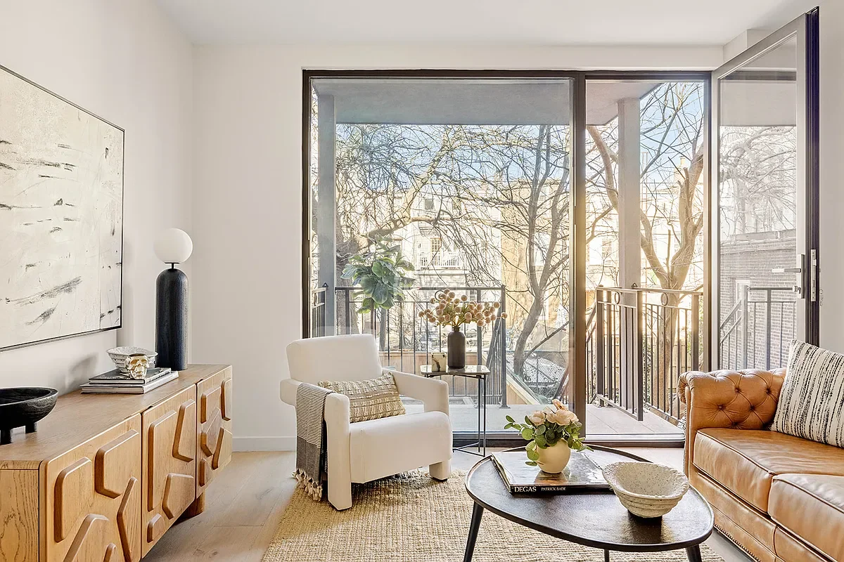 Living room with large sliding glass doors leading to a balcony, a tan leather sofa, a white armchair with a cushion, a wooden sideboard with decorative objects, a round coffee table with flowers, and a potted plant on the balcony.