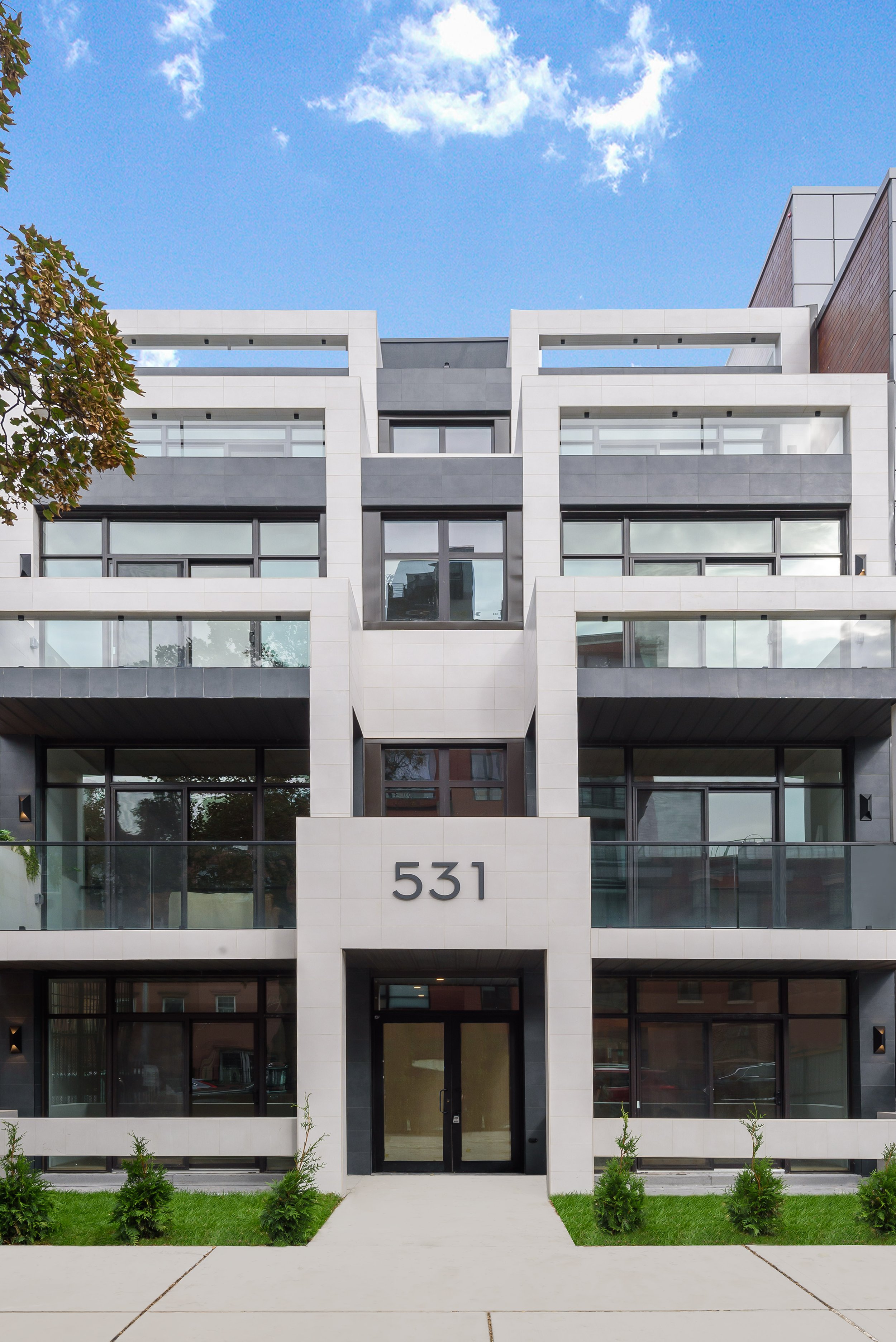 Modern multi-story residential building with white and gray exterior, large windows, small landscaped garden with green bushes in front. This is 355 Classon Avenue, a building developed by Ten Talents Management.