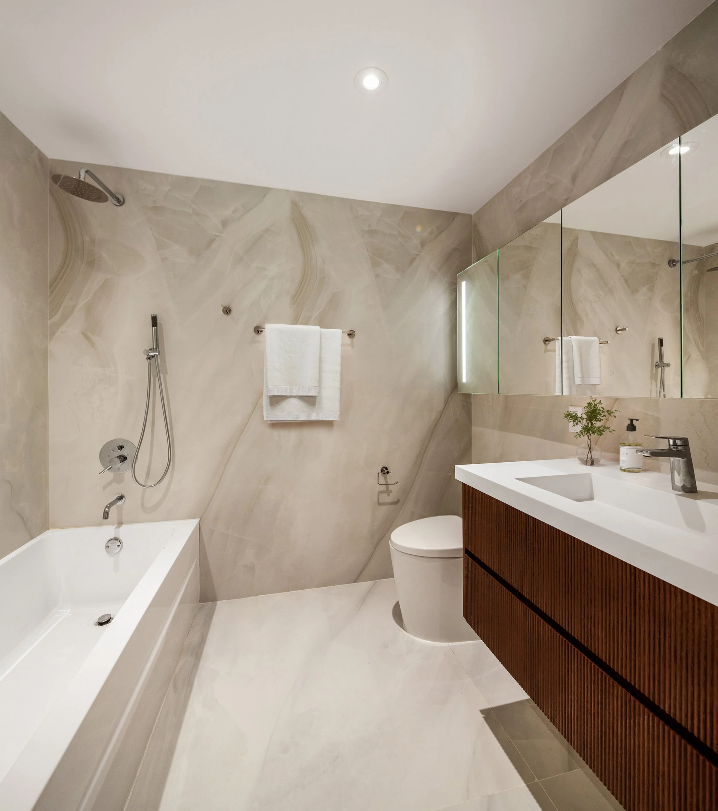 Modern bathroom featuring a bathtub with a glass shower enclosure, beige marble wall tiles, a white sink with a wooden cabinet, a toilet, a soap dispenser, and a potted plant, illuminated by a ceiling light. Developed by Ten Talents Management.