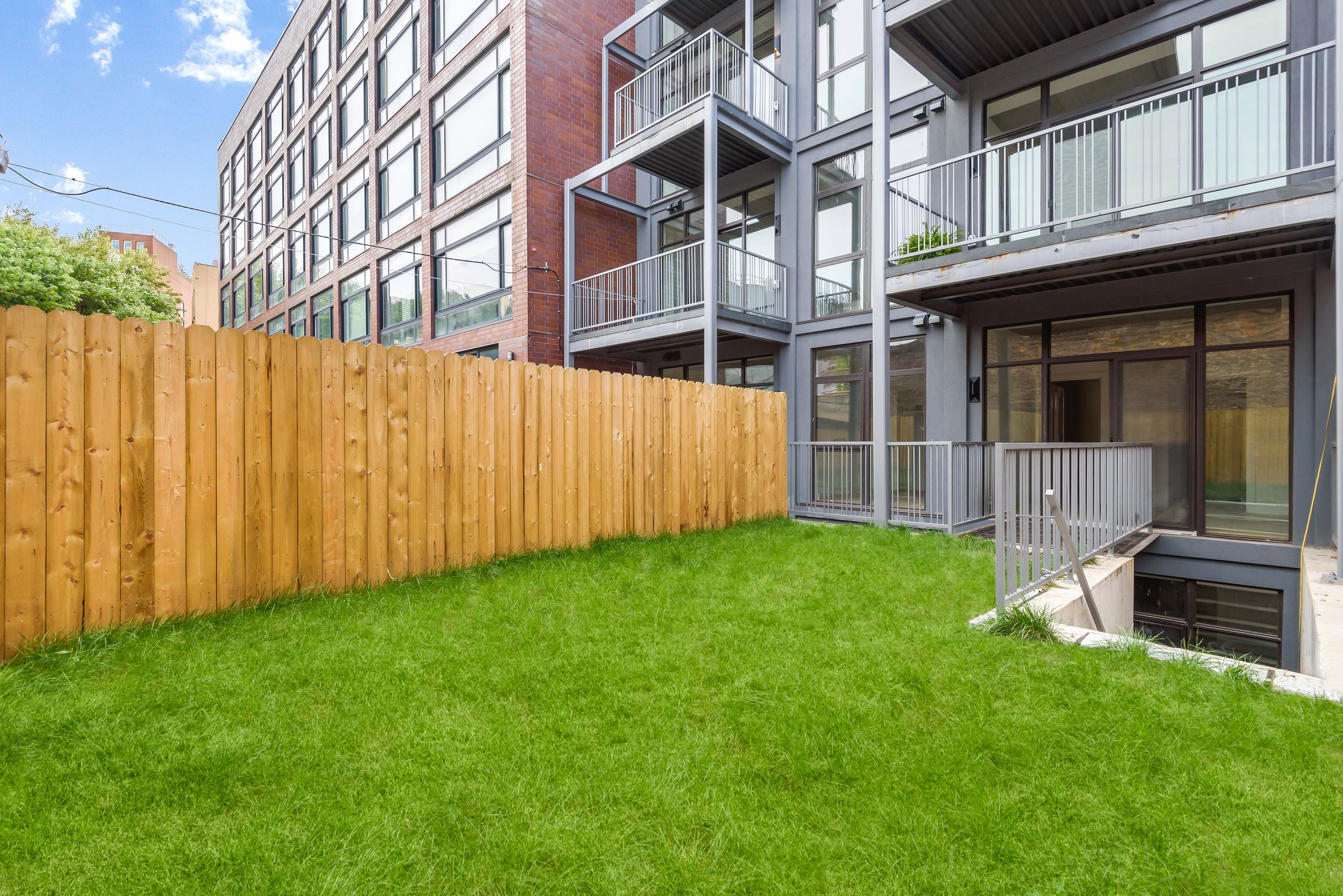 View of a modern apartment building with glass balconies and a wooden fence, green lawn, and clear blue sky. Developed by Ten Talents Management.