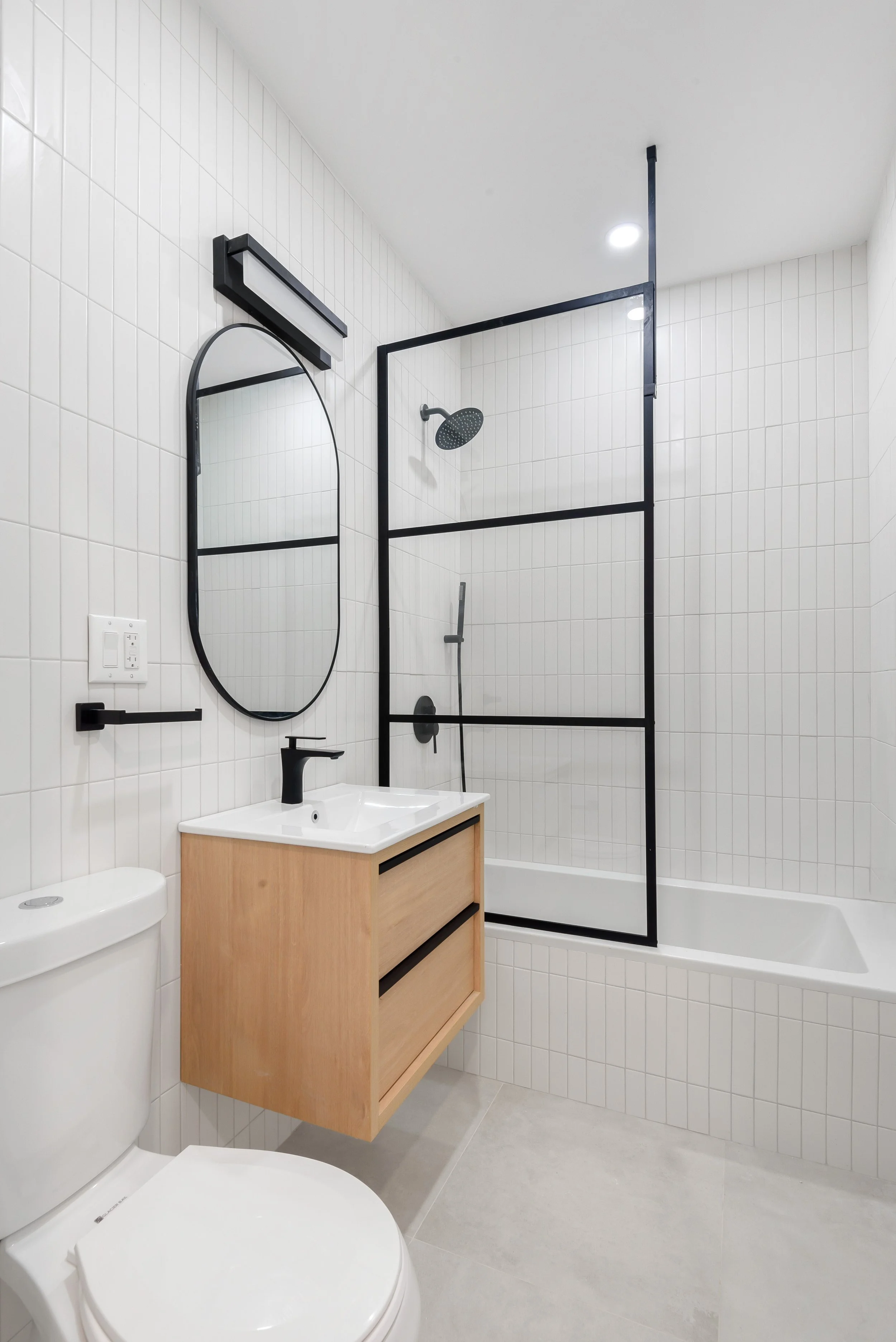 Modern bathroom with white tiled walls, a black framed glass shower enclosure, a wall-mounted mirror, a small wooden vanity with a white sink, a black faucet, and a toilet. Developed by Ten Talents Management.