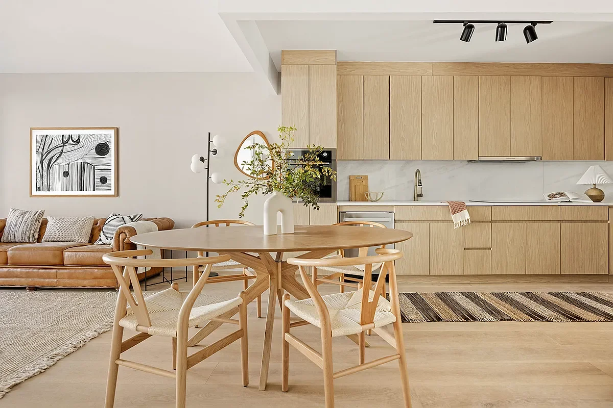 Modern open-concept living room and kitchen with light wood furniture, a tan leather sofa, black and white artwork, a round dining table with four chairs, and minimal decor. Developed by Ten Talents Management.
