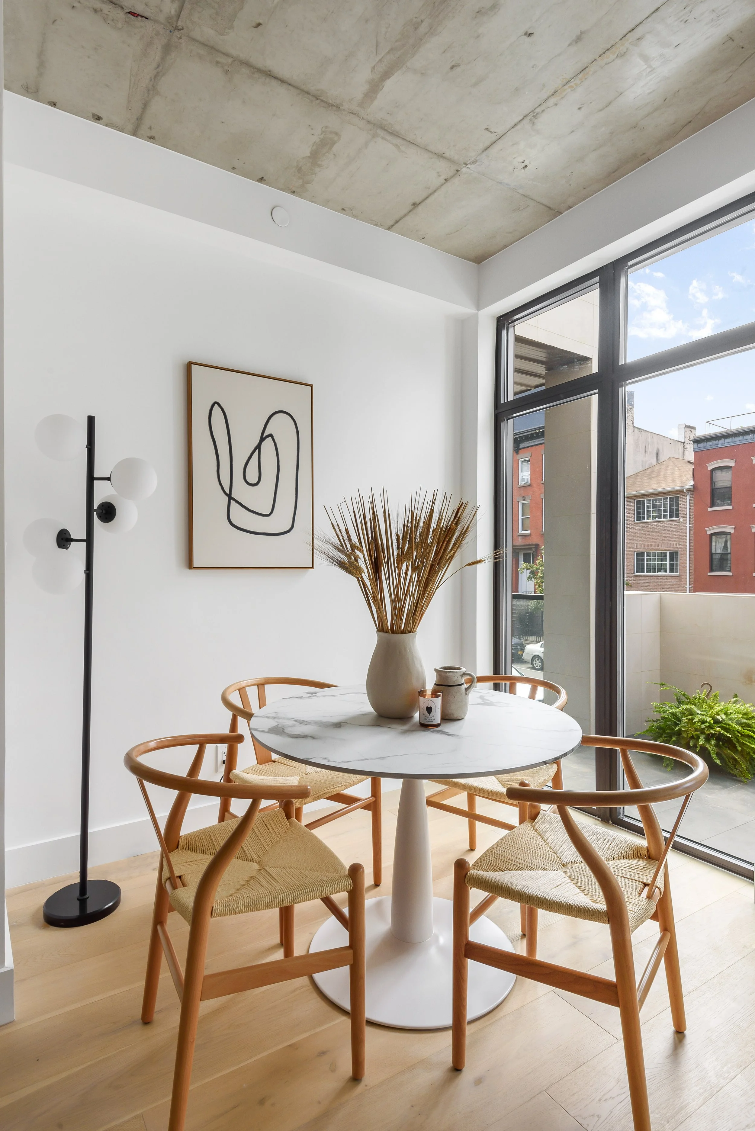A minimalist dining area with a round marble-top table, four wooden chairs with woven seats, a framed abstract line art on the white wall, and a large window with a city view of brick buildings and blue sky. Developed by Ten Talents Management.