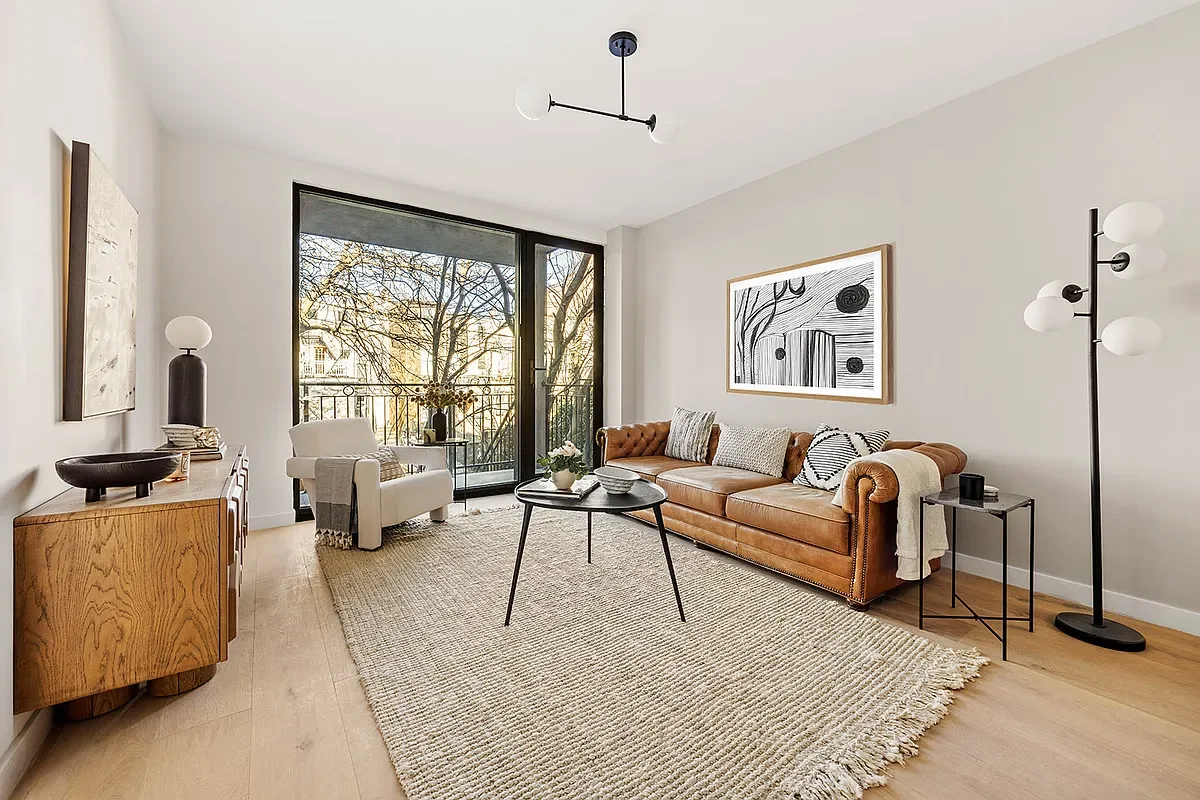 Modern living room with a leather sofa, white armchair, wooden sideboard, floor lamp, and artwork. Large glass sliding door leads to a balcony with trees outside. Developed by Ten Talents Management.