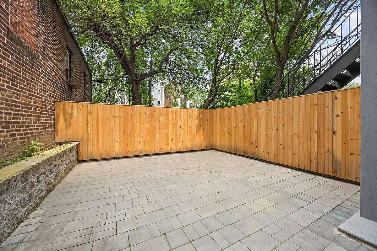 Empty backyard patio with gray paving stones, surrounded by a wooden privacy fence, a brick wall on the left, tall trees, and a metal staircase on the right. Developed by Ten Talents Management.